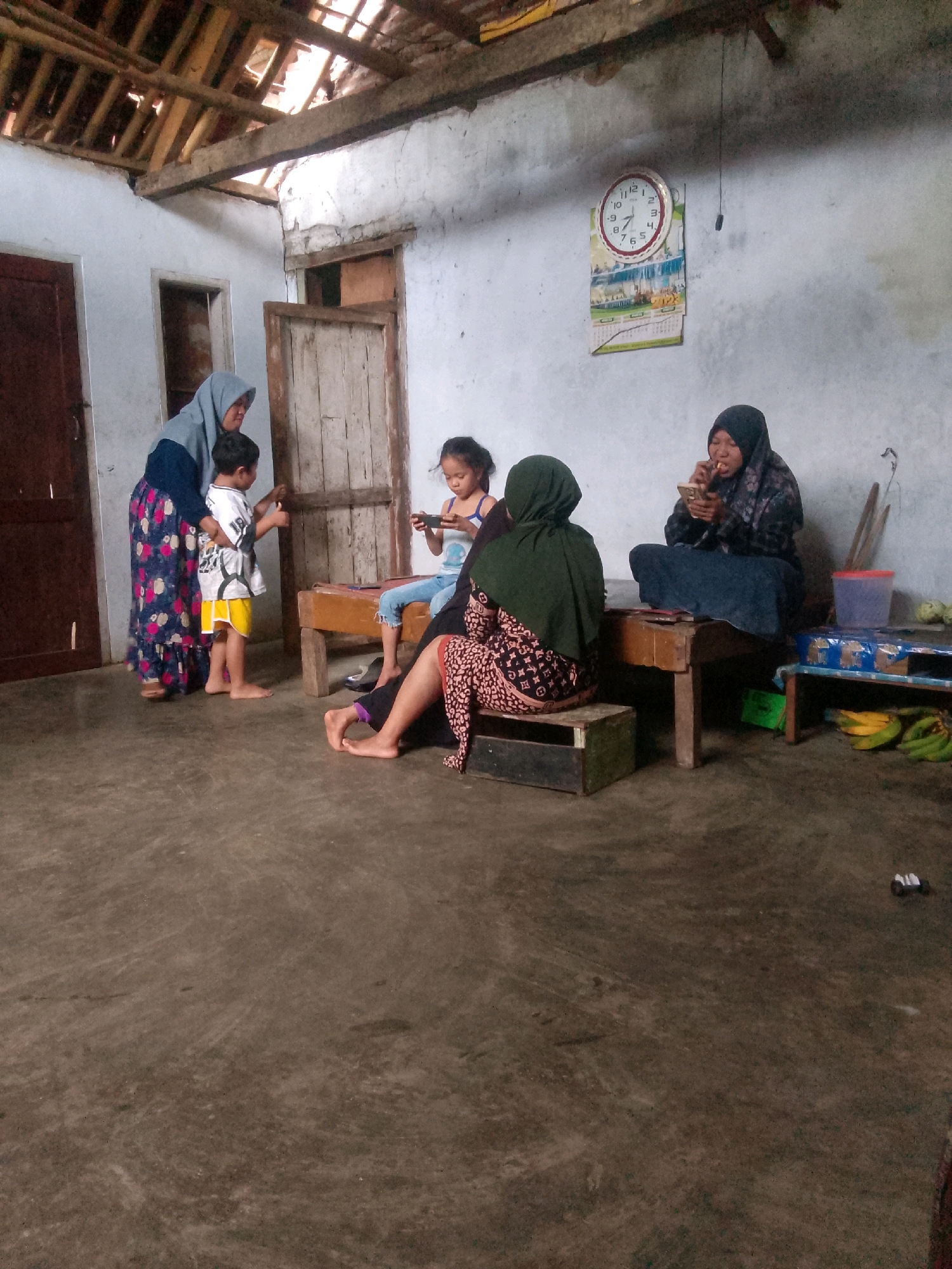 In a dimly lit, rustic interior, possibly a rural Indonesian home in Krucil, Indonesia, four individuals are present. On the left, a woman wearing a grey hijab and a floral patterned dress stands beside a young boy dressed in a white t-shirt and yellow shorts. The boy appears to be looking at something in his hand. In the center, a young girl in a blue top and jeans is seated, looking at a mobile phone. To the right, a woman wearing a black hijab and patterned top is seated and eating from a mobile phone. She is looking down at the phone while eating. In the background, a clock hangs on a white wall, next to a calendar displaying the year 2022. The room has a rough concrete floor and a wooden ceiling with exposed beams. There are simple wooden benches or tables, and some fruit, including bananas, are visible on a shelf in the background. The overall impression is of a casual, domestic scene in a humble dwelling.