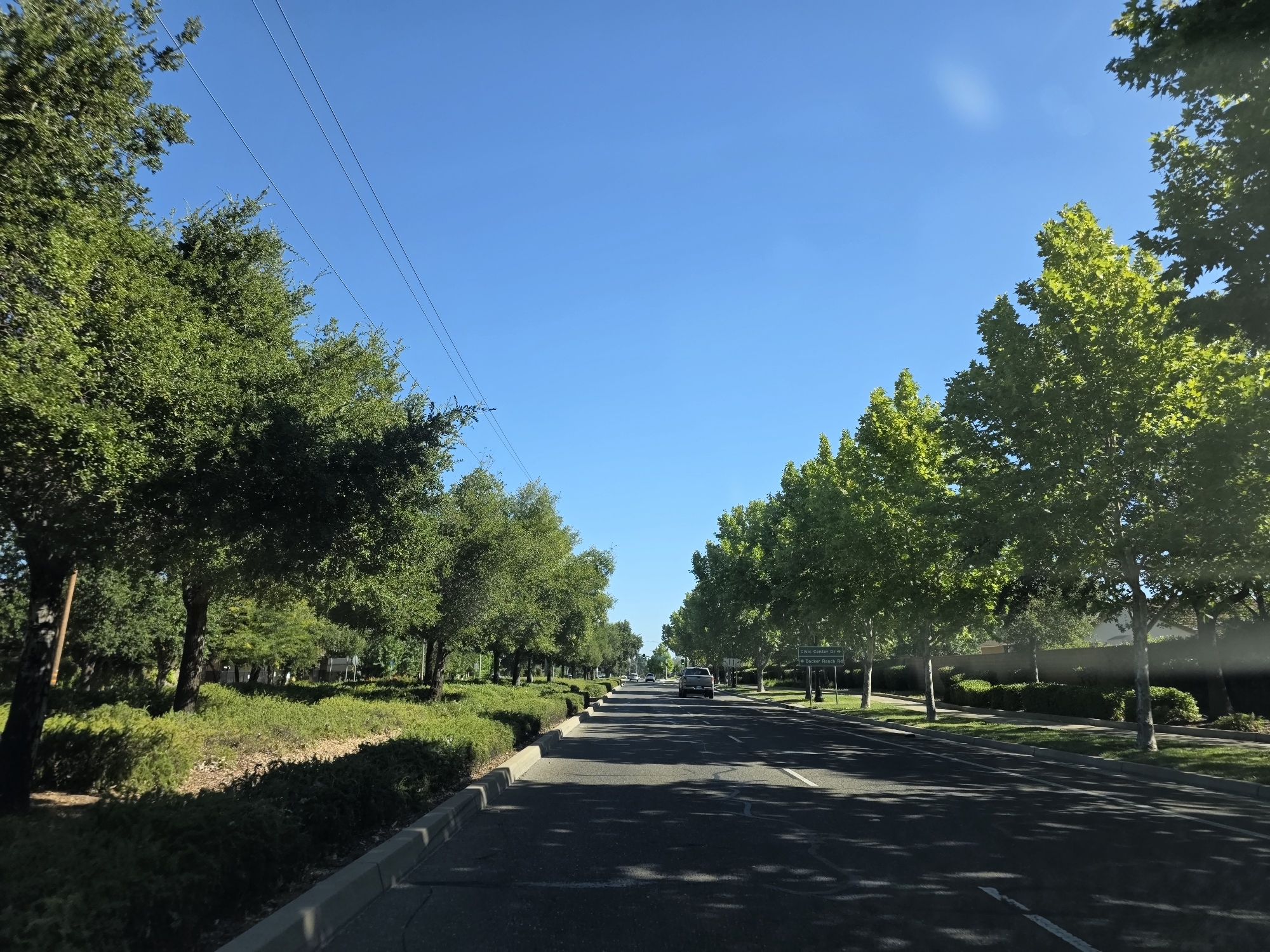 This is a landscape photograph taken from a moving vehicle, looking down a tree-lined street. The scene depicts a suburban or residential area with lush green trees lining both sides of the asphalt road. The trees are full of vibrant green leaves, suggesting a temperate climate during a growing season.
In the foreground, the road surface is visible, with white lane markings indicating a two-lane road. Shadows of the trees are cast across the road, hinting at a sunny day with the sun positioned to the side. The curbs are clearly defined, and a strip of green landscaping with bushes and smaller trees separates the road from the residential properties beyond.
Further down the road, several vehicles are visible, including a silver SUV in the middle distance. In the background, houses and other structures can be seen, partially obscured by the trees and foliage. A street sign is visible on the right side of the road, indicating directions to "Civic Center Dr" and "<- Backer Ranch Rd."
The sky is a clear, bright blue, with no clouds visible. This, along with the strong shadows, indicates that it is daytime, likely mid-morning or early afternoon. The overall impression is one of a peaceful, well-maintained suburban environment on a pleasant day. The camera appears to have a slight tilt upwards, capturing the tops of the trees against the expansive blue sky. The image has a bright and clean aesthetic, with good natural lighting.