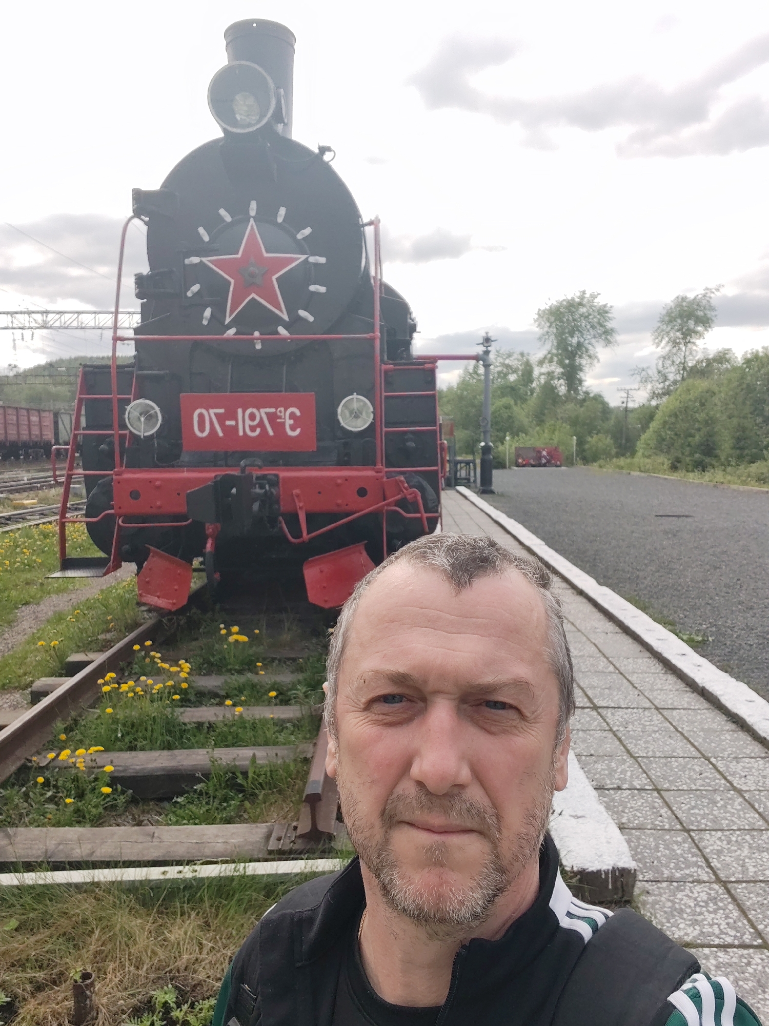 A man is captured in a selfie, positioned in the foreground with a large black steam locomotive behind him to the left. The man, with grey hair and a short beard, wears a dark jacket with white and green stripes and looks directly at the camera with a neutral expression.
The imposing steam locomotive, likely a historical model, dominates the background. Its front is black with a prominent red star, outlined in white, centered on the boiler door. Below the star, a red rectangular sign displays white, mirrored Cyrillic text, which appears to read "ЭГ-1ЭГ", identifying it as an Эг-class steam engine. The locomotive also features large headlights and red lower elements.
The setting is an outdoor railway environment, possibly a museum or a quiet station. Train tracks with wooden sleepers, partially covered in grass and yellow dandelions, run in front of and alongside the locomotive. To the right, a paved platform with a white kerb stretches into the distance. In the mid-ground, more train tracks and cargo wagons are visible. The background features trees under an overcast sky, indicating it is daytime with cloudy weather. The scene is located in Medvezhyegorsk, Russia.