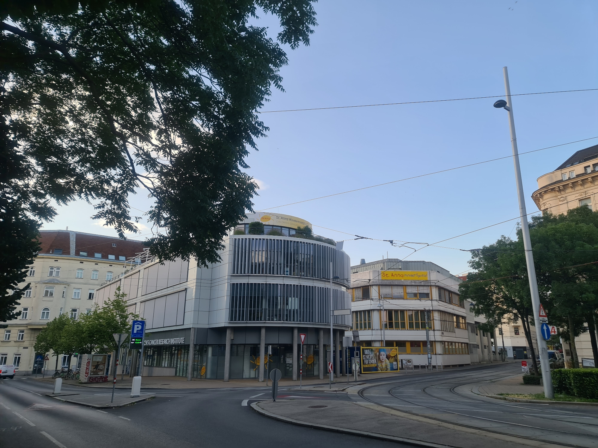 This is a daytime exterior, eye-level, wide-angle shot of a street scene in Vienna, Austria, featuring modern and older architecture, trees, and a clear blue sky. In the foreground, a paved street curves to the right, with tram tracks running along its surface. On the left, a light-colored, multi-story building with numerous windows is visible. Adjacent to it, a contemporary, curved building with vertical silver louvers and large glass windows dominates the midground. The name "SNS CANCER RESEARCH INSTITUTE" is emblazoned on its facade. To the right of this building, another yellow and white structure with a similar architectural style features prominent signage that reads "St. Anna Kinderspital" (St. Anna Children's Hospital). Several mature trees with green foliage are scattered throughout the scene, casting shadows on the ground. A tall, slender lamppost stands on the right side of the street, with overhead wires crossing the sky. Traffic signs, including a blue parking sign and a red and white yield sign, are present near the intersection. The sky is clear and bright, with a few faint clouds and distant birds visible. No people are clearly discernible in the image.