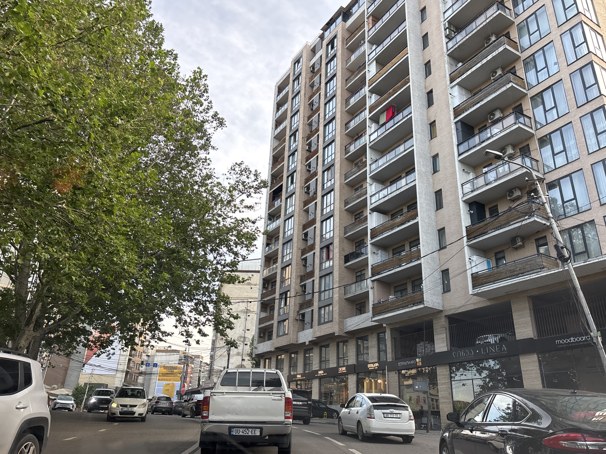 A street scene in Tbilisi, Georgia, shows a multistory apartment building with balconies and many windows, flanked by a large tree on the left and a streetlight on the right. Several cars are on the road, including a white pickup truck in the foreground. Shops with signs are visible at the base of the apartment building. The sky is overcast.