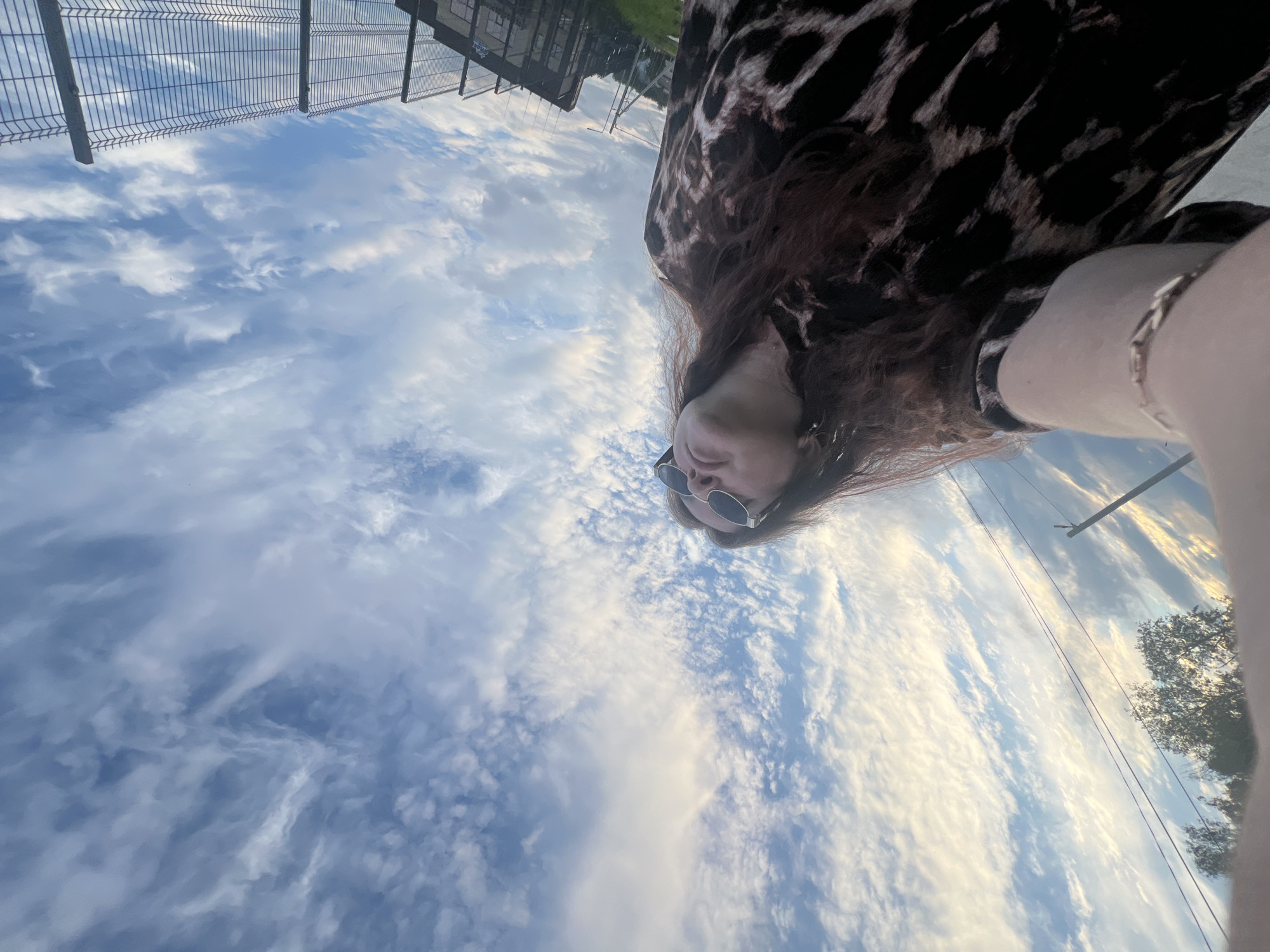 The image is a low-angle, wide shot selfie taken outdoors, likely in Noviy, Russia, during daylight hours under a sky filled with wispy white and blue clouds. In the foreground, a person's arm and shoulder are visible, wearing a bracelet. The person is also wearing a dark garment with a brown and black leopard print. Their face, partially obscured by sunglasses, is turned upwards towards the sky. In the background, a tall chain-link fence with wooden posts and a darker metal structure is visible on the right, suggesting an outdoor facility or enclosure. Utility poles and wires are present in the middle ground, along with some greenery, possibly trees or grass, and a hint of buildings. The overall scene captures a moment of leisure or observation of the sky.