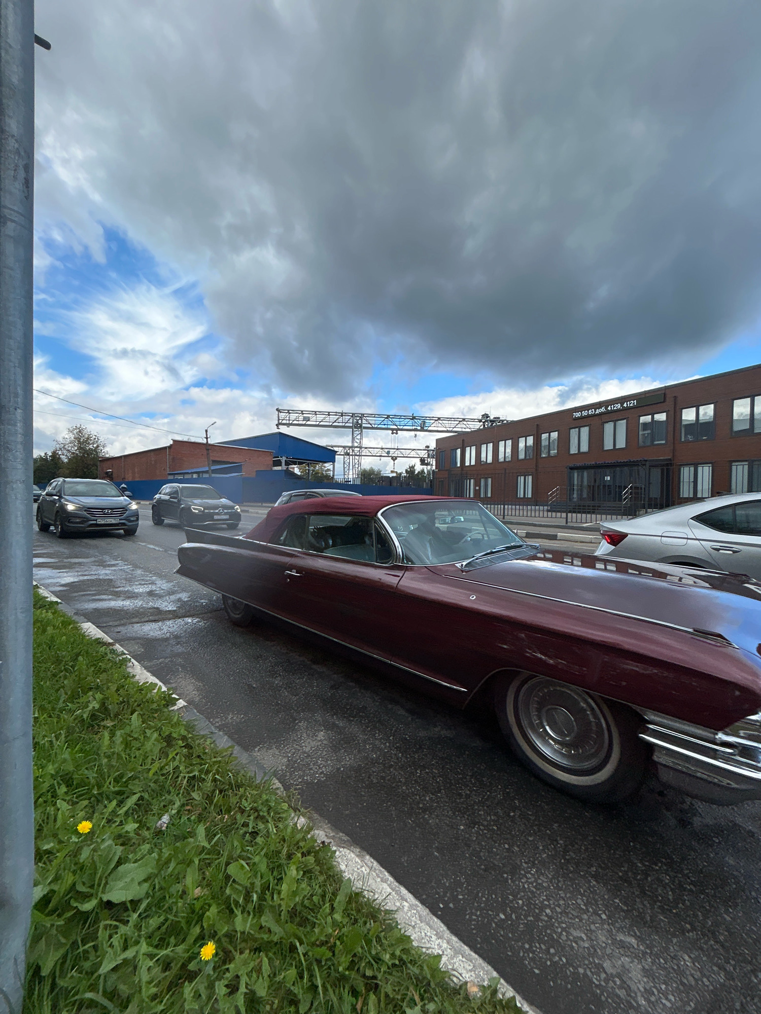 A dark red, vintage convertible Cadillac is parked alongside a wet, asphalt road in Ivanteyevka, Russia. The car is positioned at an angle, facing away from the viewer and towards the right side of the frame. In the background, several modern cars are driving on the road, including a dark grey SUV and a silver sedan. Behind the road, a brick building with numerous windows and an illuminated sign displaying "700 50 63 доб. 4129, 4121" is visible. Overhead, the sky is dramatic with a mix of grey and blue clouds. To the left of the road, a patch of green grass with scattered yellow dandelions and a metal pole can be observed. The overall scene suggests a cloudy day with recent rain.