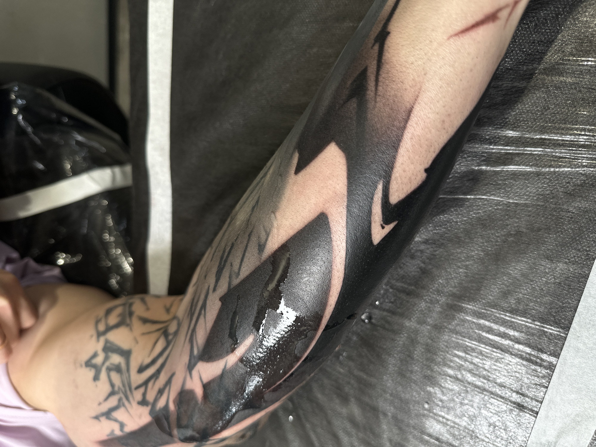 The image shows a person's arm with a large, black tattoo on the forearm. The tattoo design appears to be abstract, with sharp edges and shading. There is also a tattoo on the upper arm. The skin around the tattoo is pale. The arm is lying on a surface covered with a crinkled, gray material. A white piece of paper is to the right of the arm, and part of a black bag and a purple garment are to the left. The location is unknown.