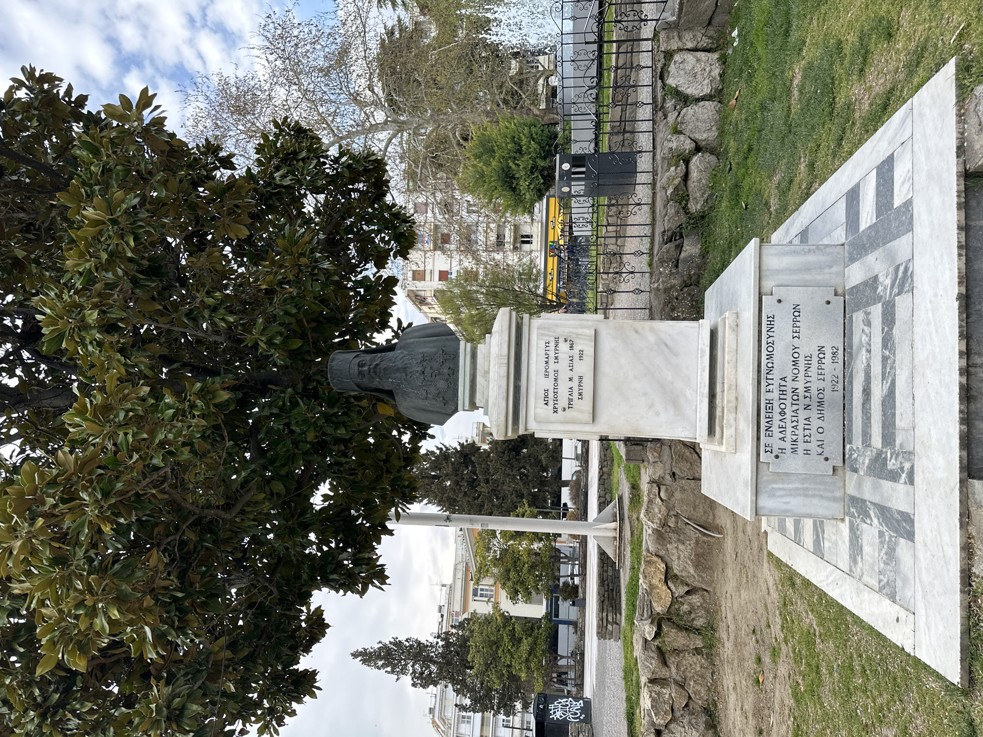 The image depicts a bust statue of Saint Chrysostomos of Smyrna in Serres, Greece. The statue is mounted on a marble pedestal inscribed with Greek text, commemorating him as a holy martyr from Smyrna, born in 1867 and martyred in 1922. The pedestal includes inscriptions indicating the memorial was erected in gratitude by the brotherhood of Asia Minor of Serres, the Hearth of New Smyrna, and the municipality of Serres. Behind the statue, there's a black iron fence, greenery, and buildings in the background. A tree's foliage partially obscures the statue. The ground around the monument consists of grass and stone elements.