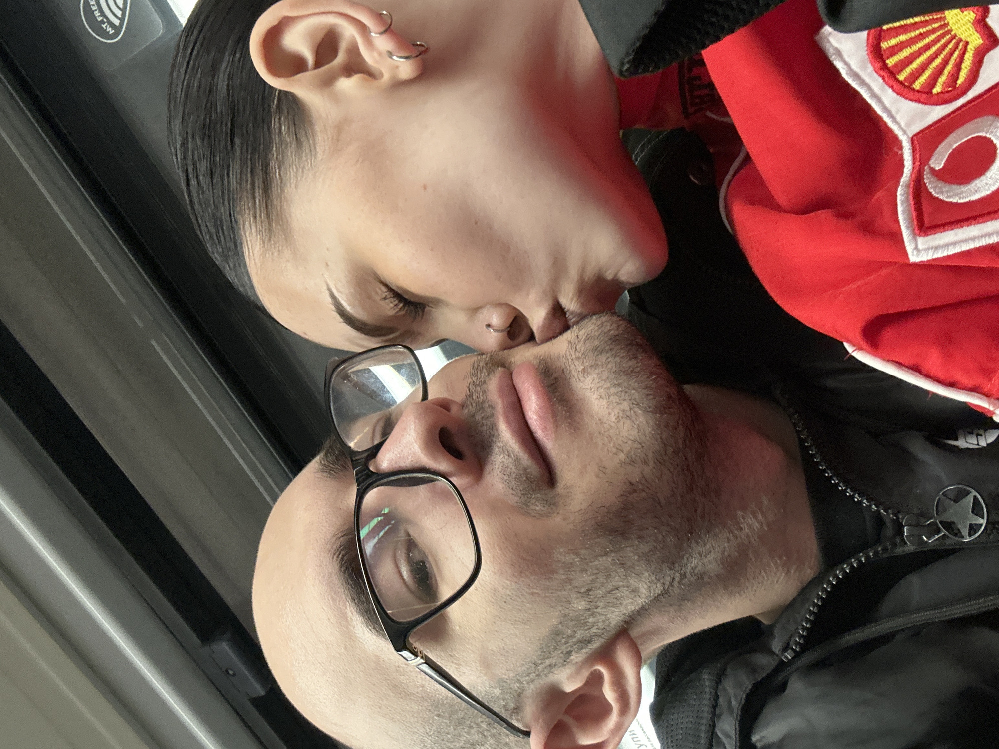 A close-up shot depicts two individuals in an intimate moment, seemingly sharing a kiss. The person on the left has dark hair, fair skin, and multiple piercings in their ear. They are wearing a red garment with patches, one featuring a Shell logo. The person on the right has a shaved head, light facial hair, and wears black-framed glasses. They are wearing a black jacket with a zipper and a star-shaped emblem.
The background is nondescript. It is not possible to determine the location as Moscow, Russia based on the image content alone.