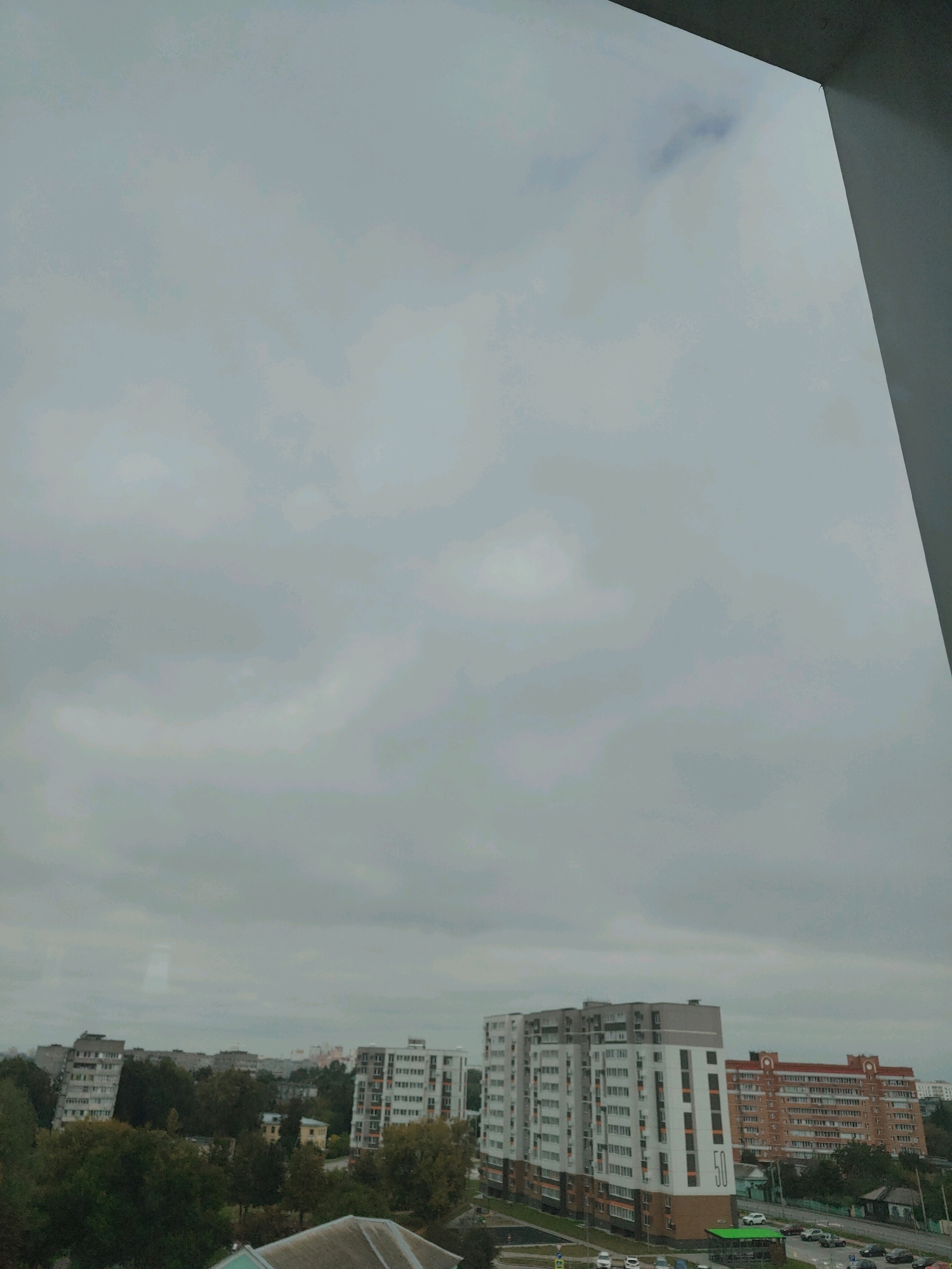 The image is a wide-angle, overcast view of an urban environment, likely Homyel, Belarus, as suggested. The sky is a uniform grey, filled with clouds. In the foreground, a sliver of a building's interior edge is visible on the right, suggesting the photo was taken from inside a room or apartment. Below the sky, a cluster of buildings forms the cityscape.
Prominent in the mid-ground are several multi-story residential buildings. One striking building on the right is tall and modern, with white and grey facades and numerous windows. It has an orange accent on its lower section and a large "50" numerical designation on its side. To its left, another building with a lighter facade and darker vertical stripes is visible. Further to the left, older, less uniform buildings are present, with some appearing to lean slightly. Dense green trees are interspersed between and around the buildings, indicating a significant presence of greenery within the urban setting.
In the lower part of the image, a street with several cars is visible, suggesting active traffic. The overall impression is of a residential district on a cloudy day. There are no visible people in the frame. The environment appears to be a typical Soviet-era planned urban area, common in many Belarusian cities.