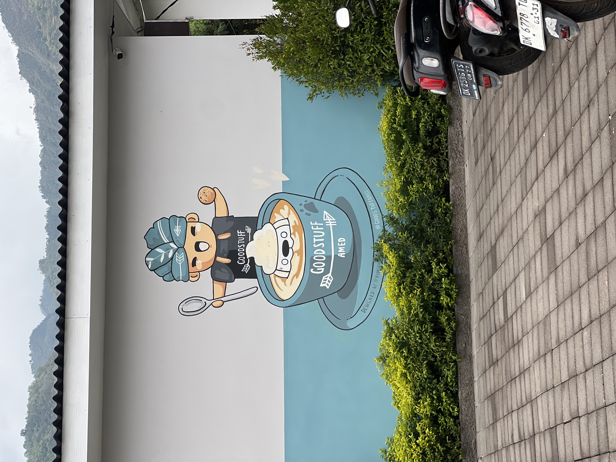 The image shows a mural on the side of a building in Abang, Indonesia. The mural depicts a cartoon character wearing a blue and green head covering, holding a spoon in one hand and a cookie in the other. The character is standing over a large blue cup filled with a light brown liquid and topped with whipped cream. A white cartoon dog's head is visible emerging from the whipped cream. The cup has the words "GOOD STUFF AMED" written on it, along with some decorative arrows and a paw print. The building's wall is painted white and light blue. In front of the wall is a row of green bushes. A paved area made of grey rectangular stones is in the foreground, with two parked scooters visible on the right side of the image. The scooters have license plates that read "DK 2536 TS" and "DK 6778 TG". In the background, a mountainous landscape is visible under a cloudy sky.