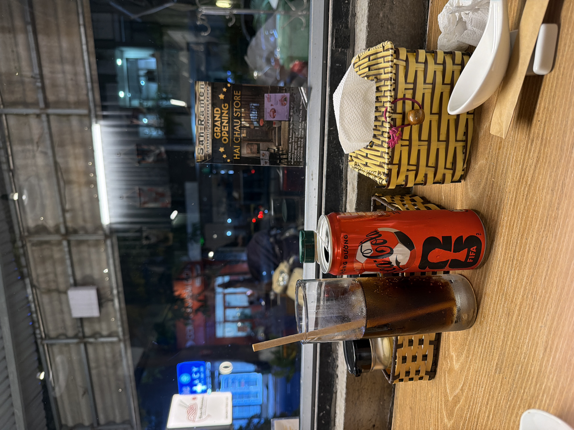 The media file captures a close-up view of a wooden tabletop next to a window, looking out onto a street at night. In the foreground, a vibrant red Coca-Cola can, clearly marked "Coca-Cola KHÔNG ĐƯỜNG" (Sugar Free) and featuring a "FIFA" logo, sits beside a glass filled with a dark carbonated beverage. A long, thin straw protrudes from the glass.
On the table, a distinctive yellow and black woven napkin holder, fashioned from a material resembling bamboo or wicker, contains white paper napkins. A white ceramic spoon and a flat, light-colored stirrer or chopstick holder are also present.
Beyond the tabletop, the window reflects the blurred lights of an urban street scene at night, indicating the late hour. Attached to the window is a poster announcing the "GRAND OPENING" of "San Ramen Authentic Japanese Ramen" at its "HAI CHAU STORE". The poster details the "STORE NAME: San Ramen Hai Chau・ラーメン", "HOTLINE: 0971 111 216", and "LOCATION: 038 Ly Thuong Kiet, Hai Chau, Da Nang". The "BUSSINESS HOUR" is listed as "11AM-11PM". The visible Vietnamese text on the Coca-Cola can and the location specified on the poster confirm the setting as Đà Nẵng, Vietnam, likely at a casual eatery.