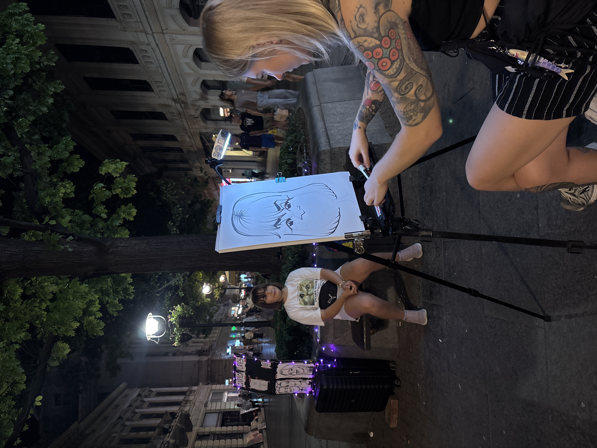 This image captures a street scene at night in Belgrade, Serbia, likely on a pedestrian street. A young girl sits on a bench to the left, posing for a caricature artist. The girl is wearing a white t-shirt with a cat graphic and a black fanny pack. She is sitting with her hands clasped in her lap. To her left is a large black suitcase with purple lights strung around it, and a sign that reads "CARICATURES" with drawings of faces.
In the foreground, the caricature artist, a woman with extensive arm tattoos, is actively drawing on a canvas on an easel. She is wearing a black top and a striped skirt. The unfinished caricature on the easel depicts a person with large eyes and distinctive hair.
The background shows a typical European street with buildings lining both sides, illuminated by streetlights and lights from shops. People are walking in the distance, suggesting a lively evening atmosphere. The presence of trees overhead indicates an urban park or a tree-lined street. The overall lighting suggests it is nighttime, with artificial lights illuminating the scene.