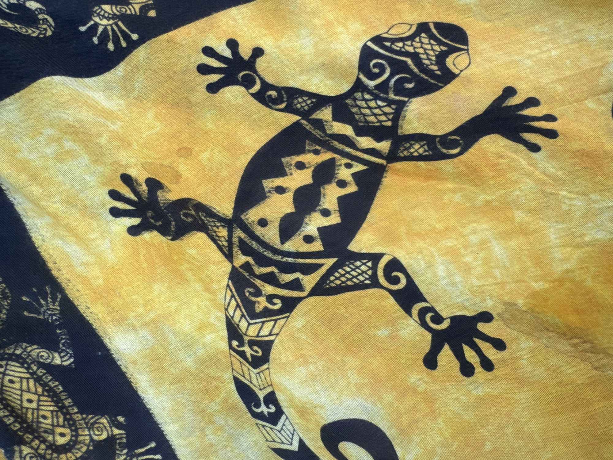 The image shows a piece of fabric with a yellow and black pattern. The primary subject is a large, stylized black gecko design in the center, adorned with various geometric and curvilinear patterns. Smaller, similar gecko designs are visible in the upper left and lower left corner of the fabric. The background is a mottled yellow color. The fabric appears to be draped or laid out, exhibiting folds and creases. There is no location information specified.