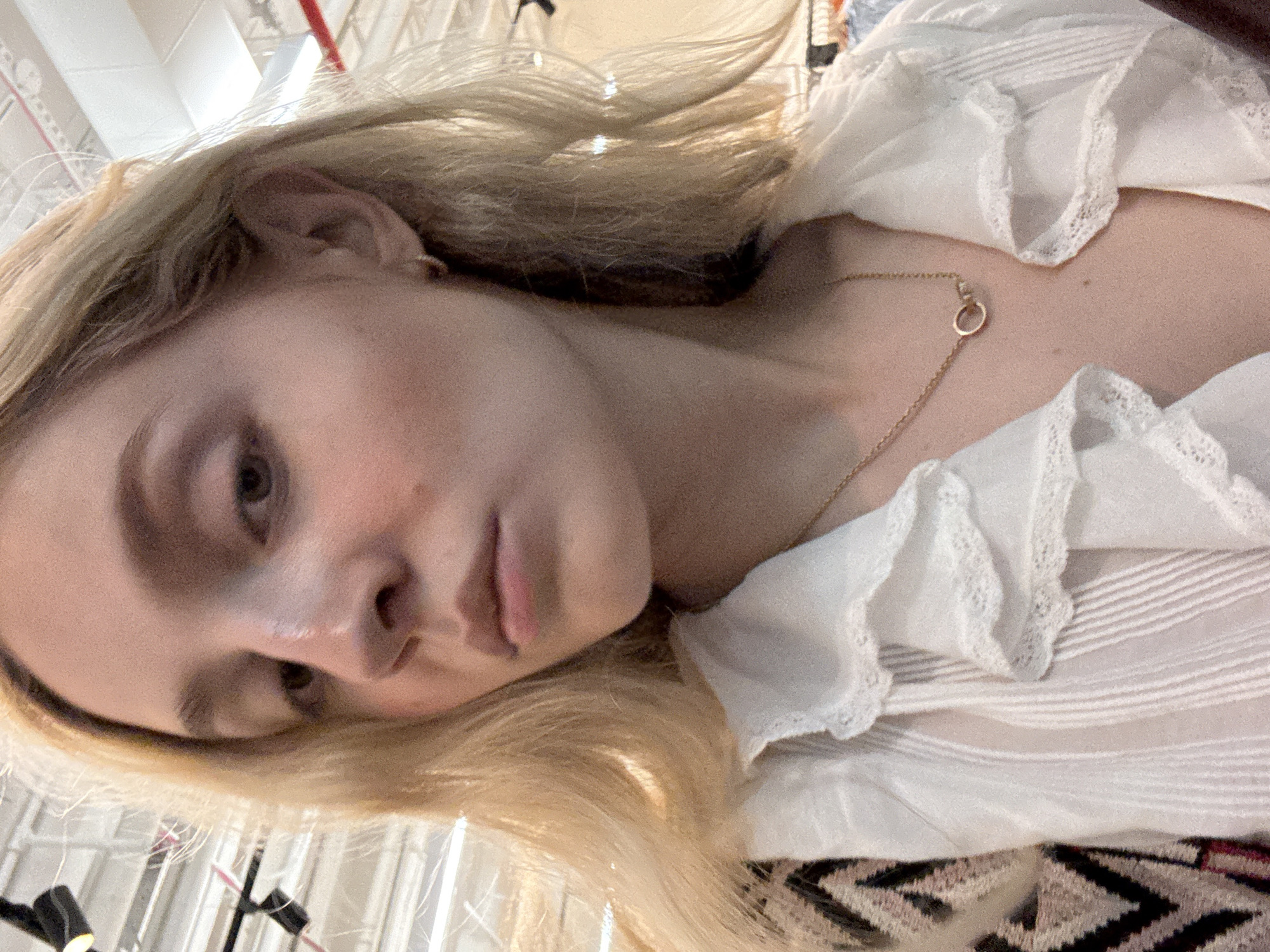 A young, fair-skinned woman with blonde hair and light makeup poses for a selfie. She is wearing a white ruffled blouse with lace trim and a gold chain necklace with a circular pendant. The background appears to be an indoor setting with fluorescent lighting and ceiling fixtures, possibly a retail store or public space in New York City, United States of America. The woman is looking down and to the side, with a neutral expression. The image is a close-up shot, focusing on her upper body and face.