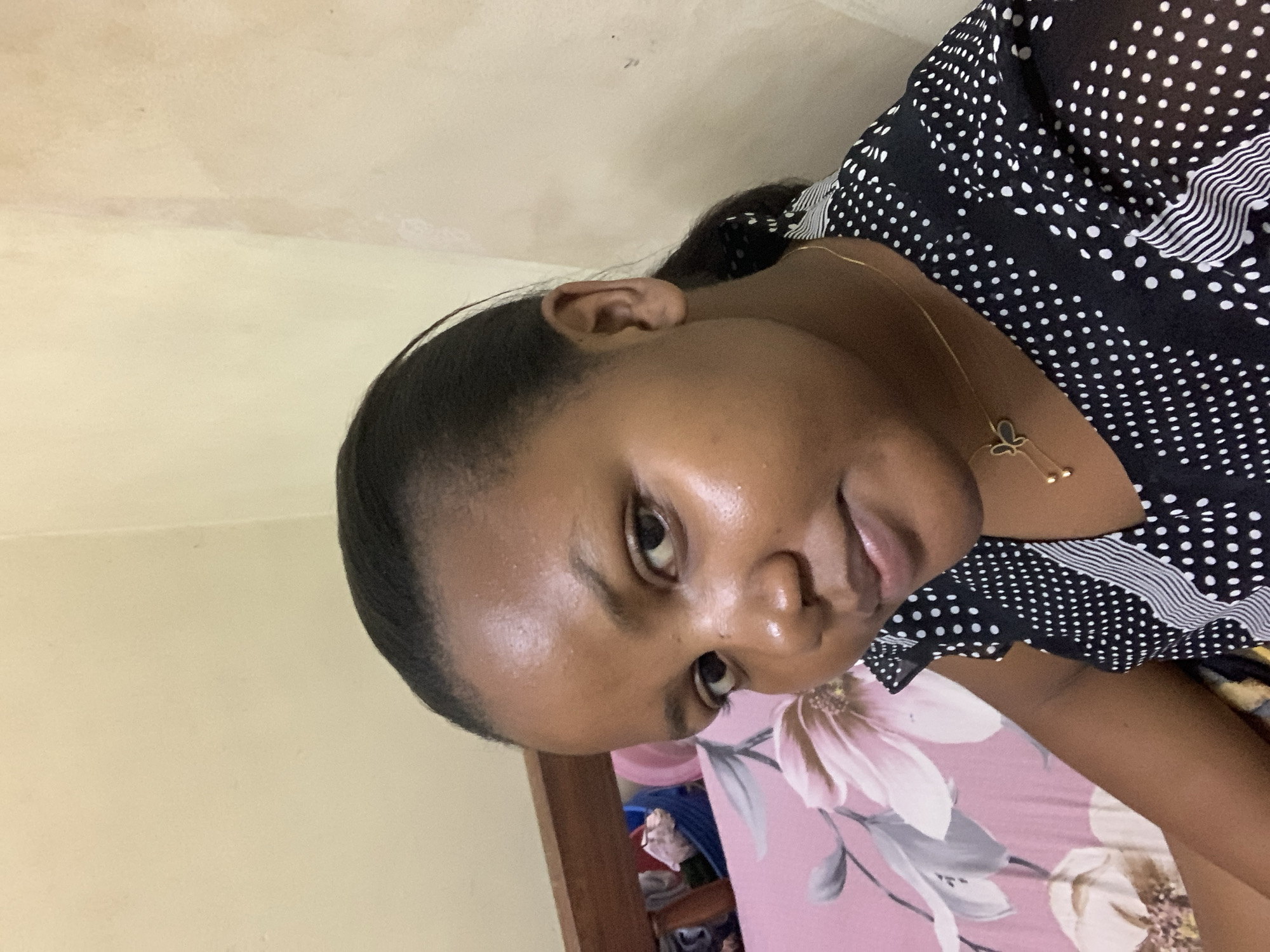 The image shows a woman lying on a pink floral patterned bed. She is wearing a black and white polka dot top. She has dark hair pulled back and is wearing a gold necklace with a pendant. The background consists of a light yellow wall. The location in the city of Dar es-Salaam, Tanzania, cannot be determined from the image.
