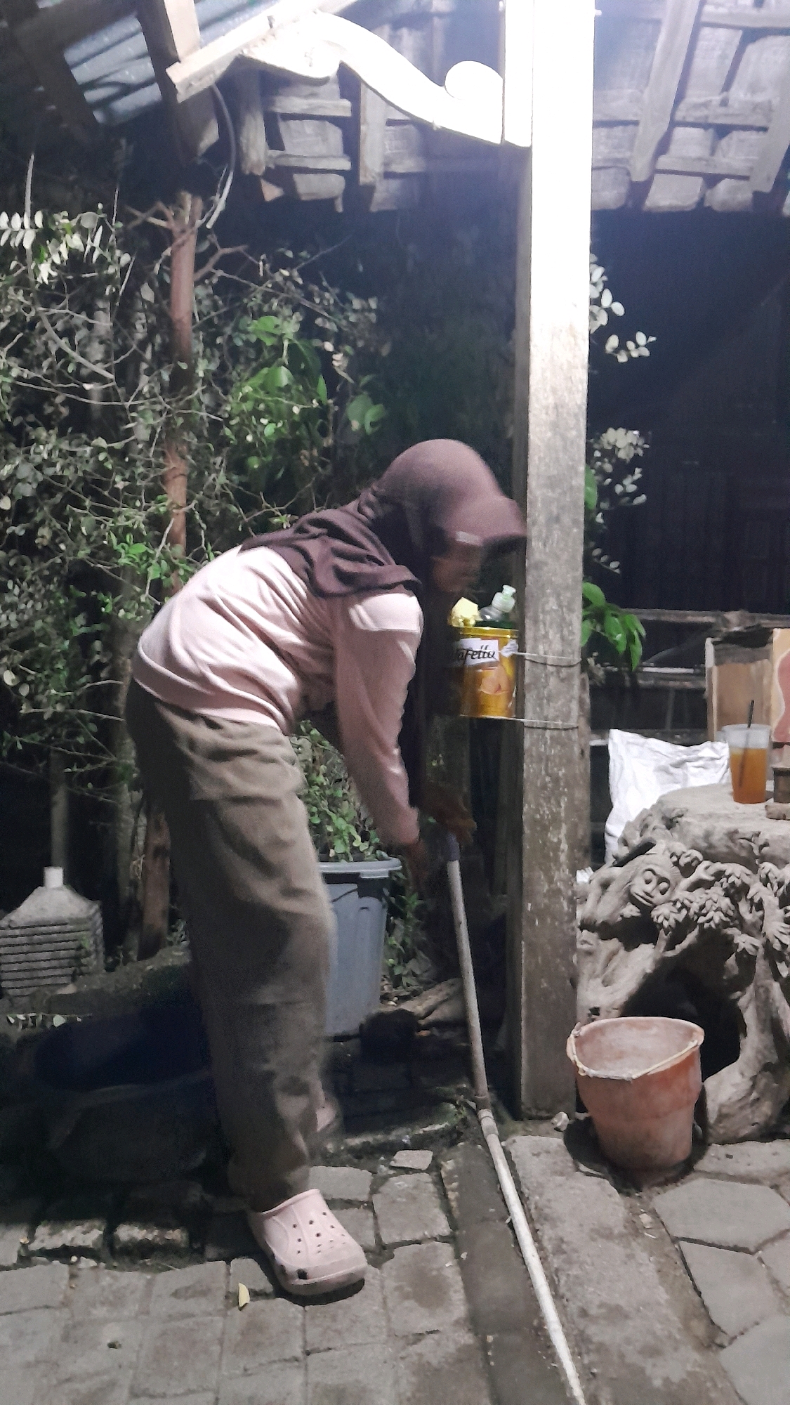 A person wearing a headscarf and a light pink shirt is sweeping the ground with a broom. The person is also wearing brown pants and light pink Croc-style shoes. They are standing on a paved area next to a wooden structure. To the right of the person, a yellow bucket with a label that says "Fettu" is hanging from a wooden post. There is also a red bucket on the ground near a carved stone sculpture. In the background, there are plants and trees. The scene appears to be outdoors, possibly a backyard or a communal area, at night or in low light. The location is in Sumberarum, Indonesia.