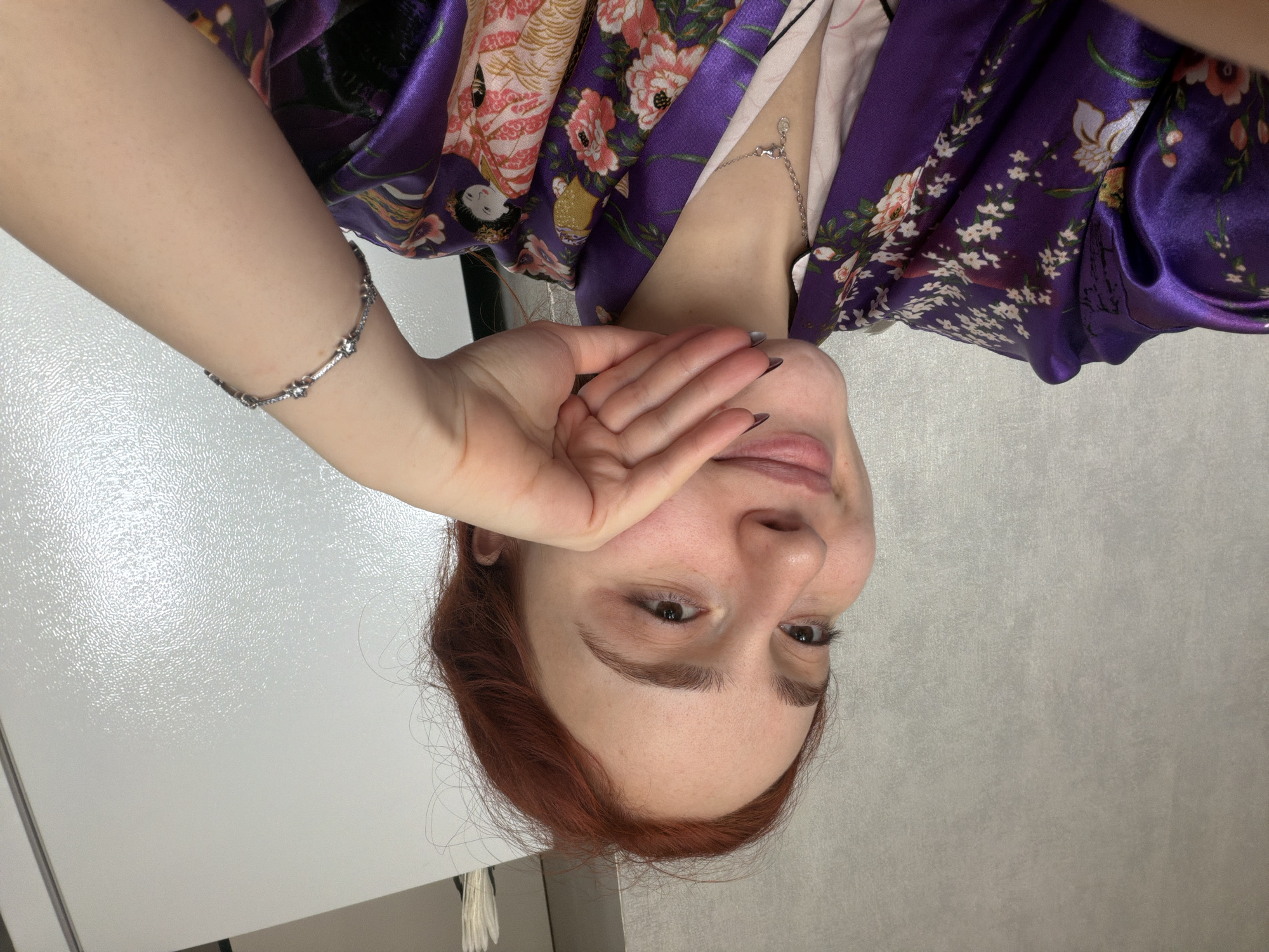 An indoor, portrait-style photograph features a young woman with reddish-brown hair and light skin, looking towards the viewer with a soft expression and a slight smile. Her left hand is raised, covering her mouth and chin, with dark nail polish visible on her fingertips. A delicate silver bracelet with small decorative elements adorns her left wrist. Around her neck, she wears a silver chain necklace with a small pendant. She is dressed in a vibrant purple kimono or robe, richly patterned with pink, white, and yellow floral motifs, and some traditional Japanese figures. The background consists of two distinct surfaces: a textured, glossy white surface, possibly a refrigerator or cabinet, on the left, and a plain, light grey wall on the right. The lighting suggests an indoor setting. No specific time of day or outdoor weather conditions are discernible, and there is no visible text.