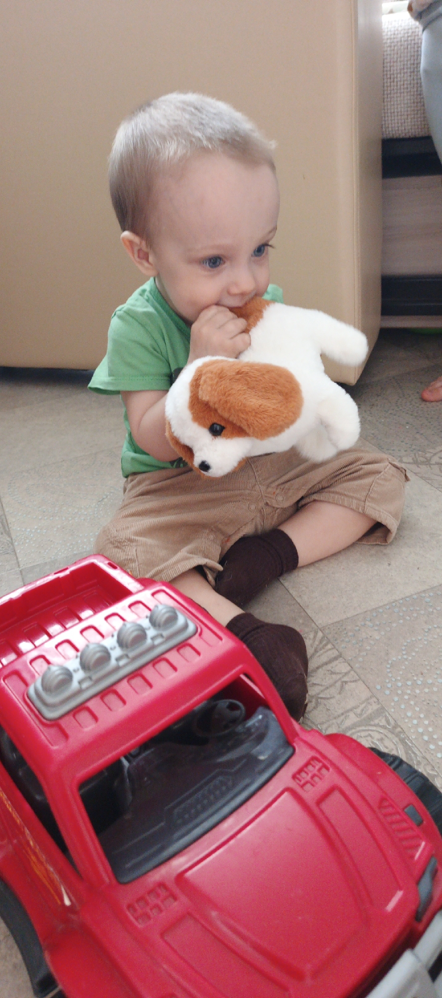 A young child is sitting on the floor next to a red toy truck, holding a stuffed dog toy in their left hand and putting their right hand into their mouth. The child has light-colored hair, blue eyes, and is wearing a green t-shirt, brown shorts, and brown socks. The stuffed dog is white with brown spots. The toy truck is large and red, with a gray rack of lights on top. The floor is tiled in a light gray pattern. A beige chair or sofa is visible in the background, along with a person's leg. The photo was taken in Chaikovsky, Russia.