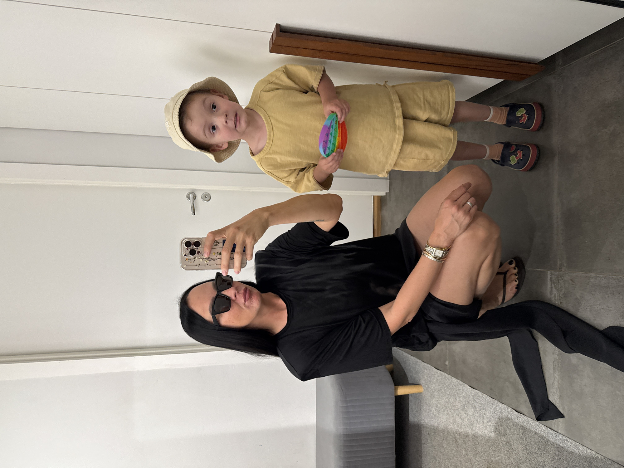 A woman and a young child are reflected in a mirror, posing for a selfie. The woman is wearing a black dress and sunglasses, and is crouched down. The child is wearing a light-colored hat and shorts, and is holding a colorful toy. The background appears to be an indoor setting, possibly a changing room or hallway, with a white door and a grey bench visible. The floor is tiled. The lighting is bright and even, suggesting an artificial light source. There is no indication of the location being in the city of Chaikovsky, Russia.
