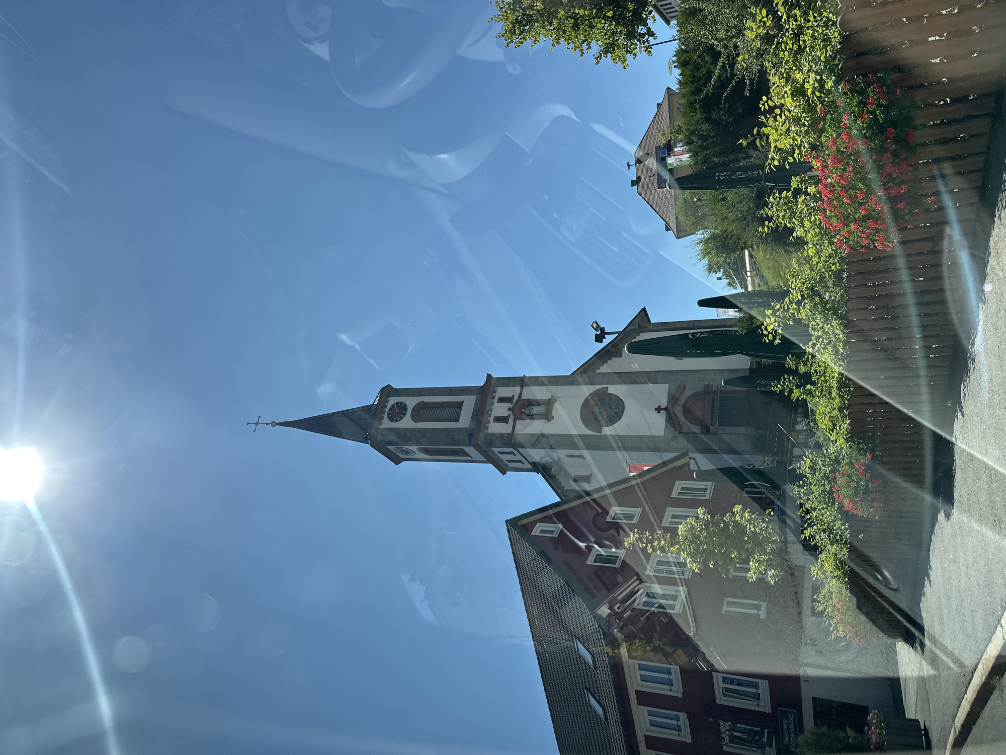 The image captures a view of the St. Lorenz Church in Schönwald im Schwarzwald, Germany. The church, with its prominent bell tower topped with a cross, is the central subject, flanked by adjacent buildings. To the left, a multi-story building with a red facade and white-framed windows is visible. Green umbrellas and lush vegetation, including red flowers, line the foreground, separated by a wooden fence. The scene is illuminated by bright sunlight, causing lens flares and reflections on what appears to be a glass surface, possibly a car window. The sky is clear and blue. The overall impression is a sunny day in a German town.