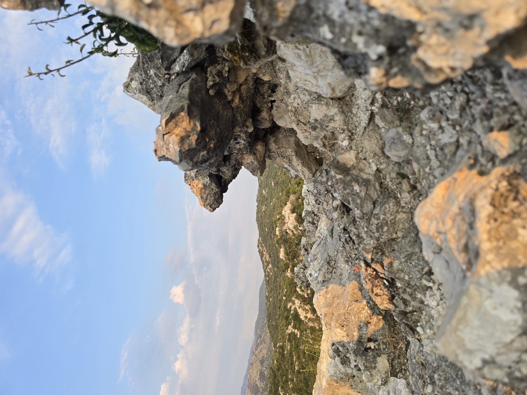 This is a close-up, low-angle shot of rough, uneven rock formations in Medun, Montenegro, likely near the fortress. The rocks in the foreground are varied in texture and color, with some appearing lighter and weathered, while others are darker and more jagged. Sunlight highlights the edges of some of the rocks, casting warm tones.

In the mid-ground and background, a lush, green, hilly landscape stretches out under a bright blue sky dotted with soft, white clouds. The terrain is covered in dense vegetation, interspersed with lighter patches that may be more rocks or sparser growth. The sky occupies the upper portion of the image, with a few delicate wisps of clouds. A few thin, green stalks with small leaves are visible growing vertically from the rocks on the right side of the frame, reaching towards the sky. The overall impression is of a natural, somewhat rugged outdoor environment.