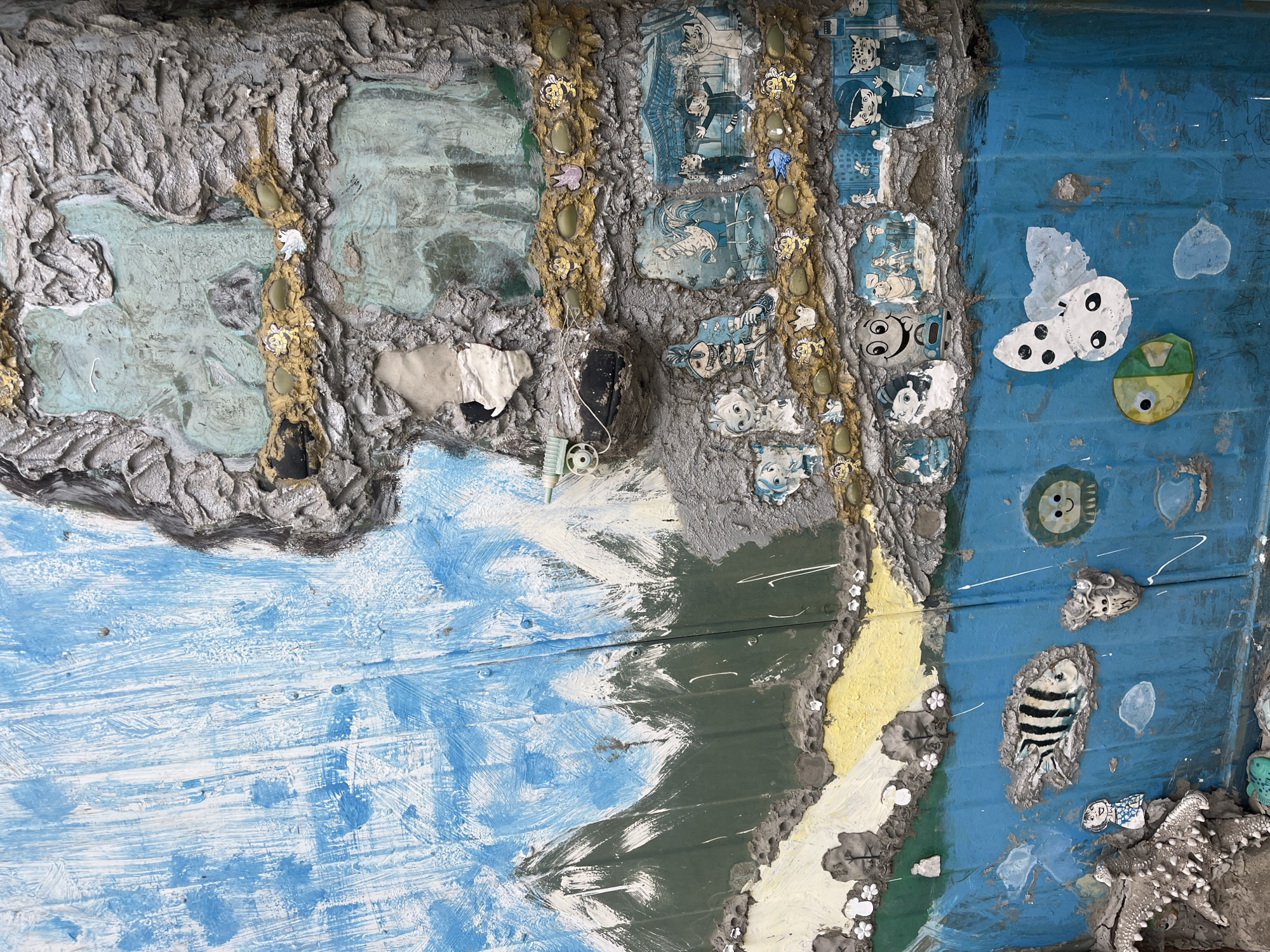 The image shows a section of what appears to be an art installation, likely part of a larger structure in Burgas, Bulgaria. The artwork incorporates various materials and techniques, including painting, sculpting with cement-like material, and the addition of found objects. The left portion features a heavily textured, gray, sculpted section with embedded fragments of fabric, plastic, and possibly broken tiles. The central area has a vertical strip of the same gray material, adorned with small, golden-painted figures, green stones, and pieces of cut-up cartoon-like images. The right side of the image displays a blue painted surface with horizontal lines, indicating either wooden planks or metal siding. This blue surface is further embellished with affixed figures of animals, including a butterfly, a fish, and various cartoon-like faces. Small shells and starfish are also embedded in the gray, textured material, suggesting a nautical theme. The overall impression is one of a whimsical and eclectic mixed-media artwork.
