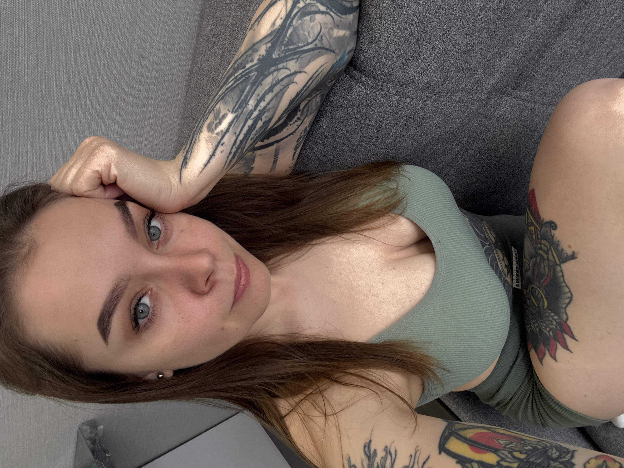 A young woman with fair skin and light brown hair is angled upward, lying on a gray sofa. Her left arm is raised, with her hand resting on her forehead. Visible tattoos adorn both arms and her right thigh. She is wearing a green, fitted top and shorts. The background includes a gray wall and a portion of a window or mirrored surface. There is no evidence in the image to indicate the specific location in the city of Gelendzhik, Russia.