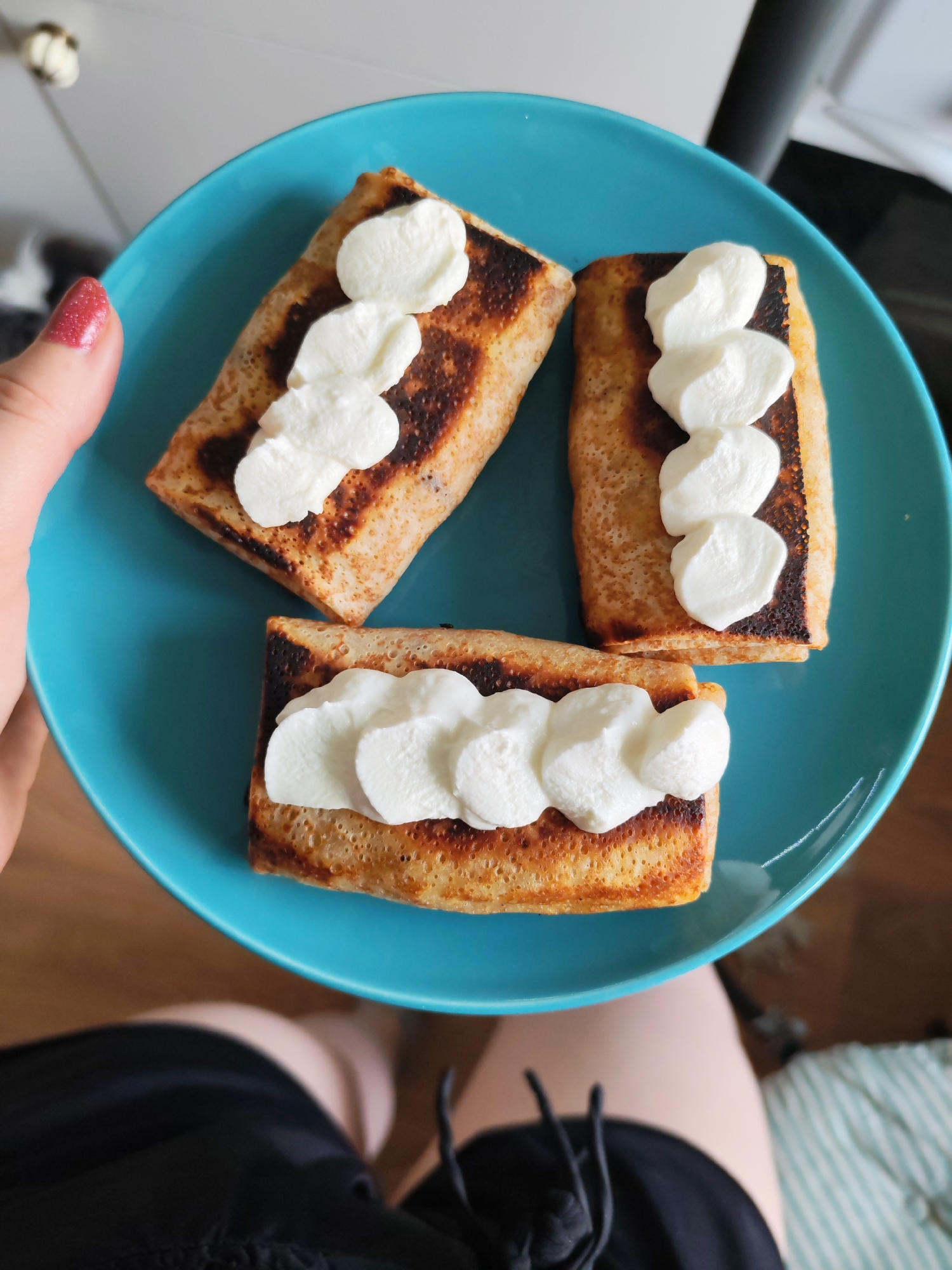 A person is holding a turquoise plate with three rolled pancakes topped with dollops of white cream. The pancakes are rectangular and slightly browned. The person has red glitter nail polish. The background includes a wooden floor, a black garment, and a glimpse of a white and grey striped fabric. The location is not discernible from the image.