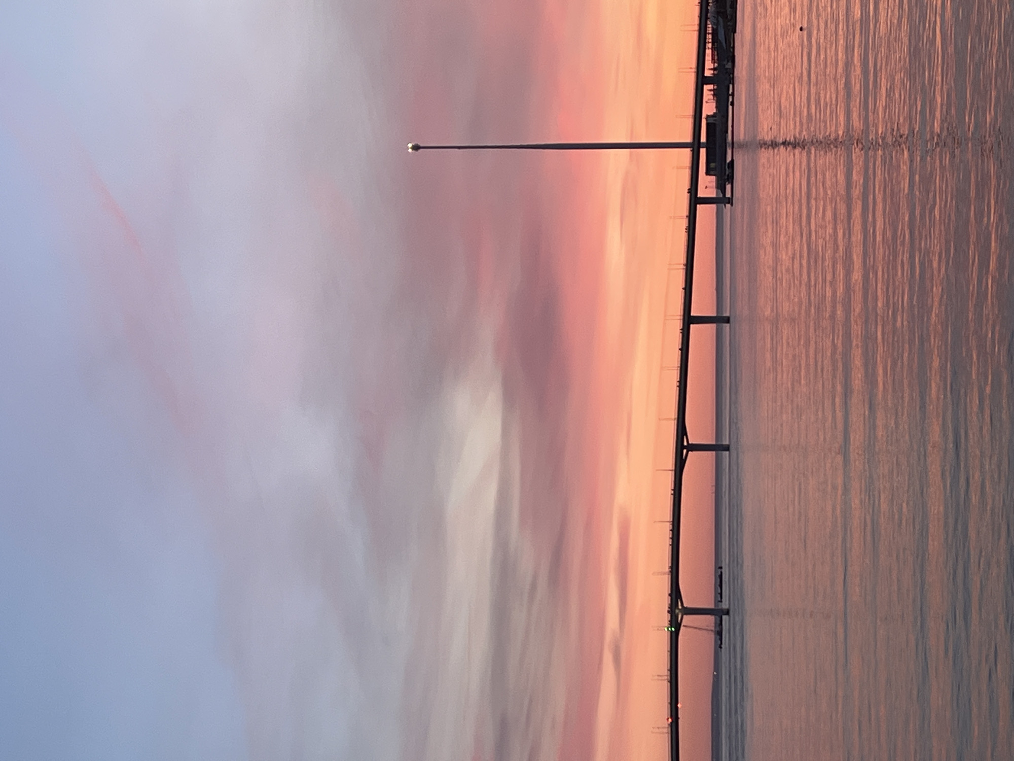 The image, oriented vertically, captures a serene sunset or sunrise over a wide body of water, likely near Saint Petersburg, Russia. A long, low-lying bridge spans horizontally across the scene, dividing the sky from its reflection on the water's surface. The sky is painted with soft, streaky clouds in gradients of pink, orange, purple, and pale blue. These warm hues are mirrored in the calm water below, creating a shimmering expanse with subtle ripples.
A tall street light pole, topped with an illuminated lamp, stands prominently on the bridge, adding a solitary point of light. The bridge structure itself is dark, appearing in silhouette against the vibrant sky and water, with multiple supports visible extending into the distance. The scene evokes a sense of tranquility and natural beauty, characteristic of twilight or dawn. There are no people, explicit activities, or visible text in the image.