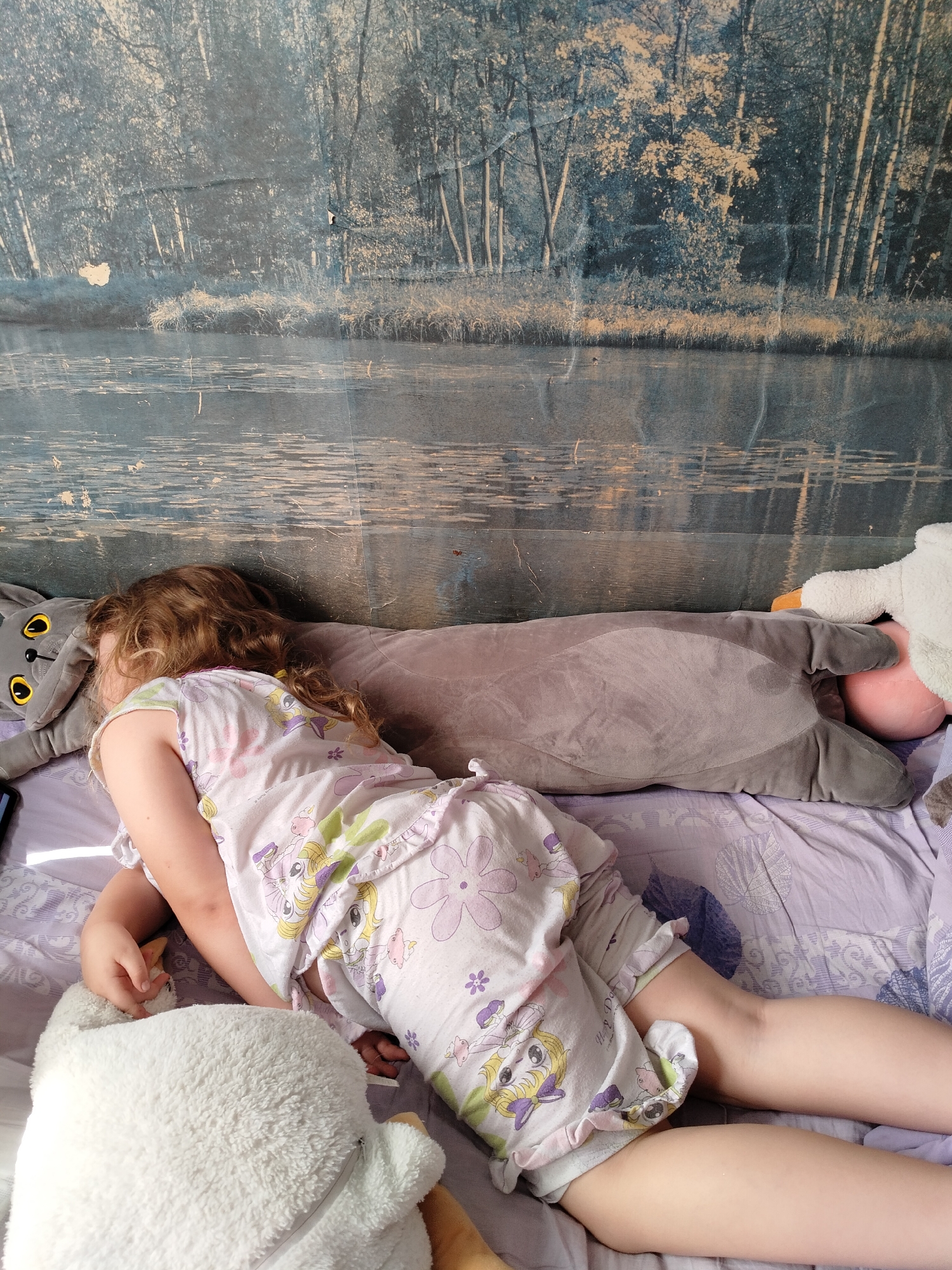 A young child with blonde hair is sleeping on a bed. The child is wearing light-colored pajamas with cartoon characters and floral patterns. The child is lying on their side, facing away from the camera, and is surrounded by several large stuffed animals, including a long gray stuffed cat and a white fluffy stuffed animal. The bed has a purple patterned sheet. In the background, a large tapestry or wallpaper depicts a serene forest scene with a body of water, characterized by muted blue and gray tones and a distressed, textured appearance. The scene appears to be indoors.