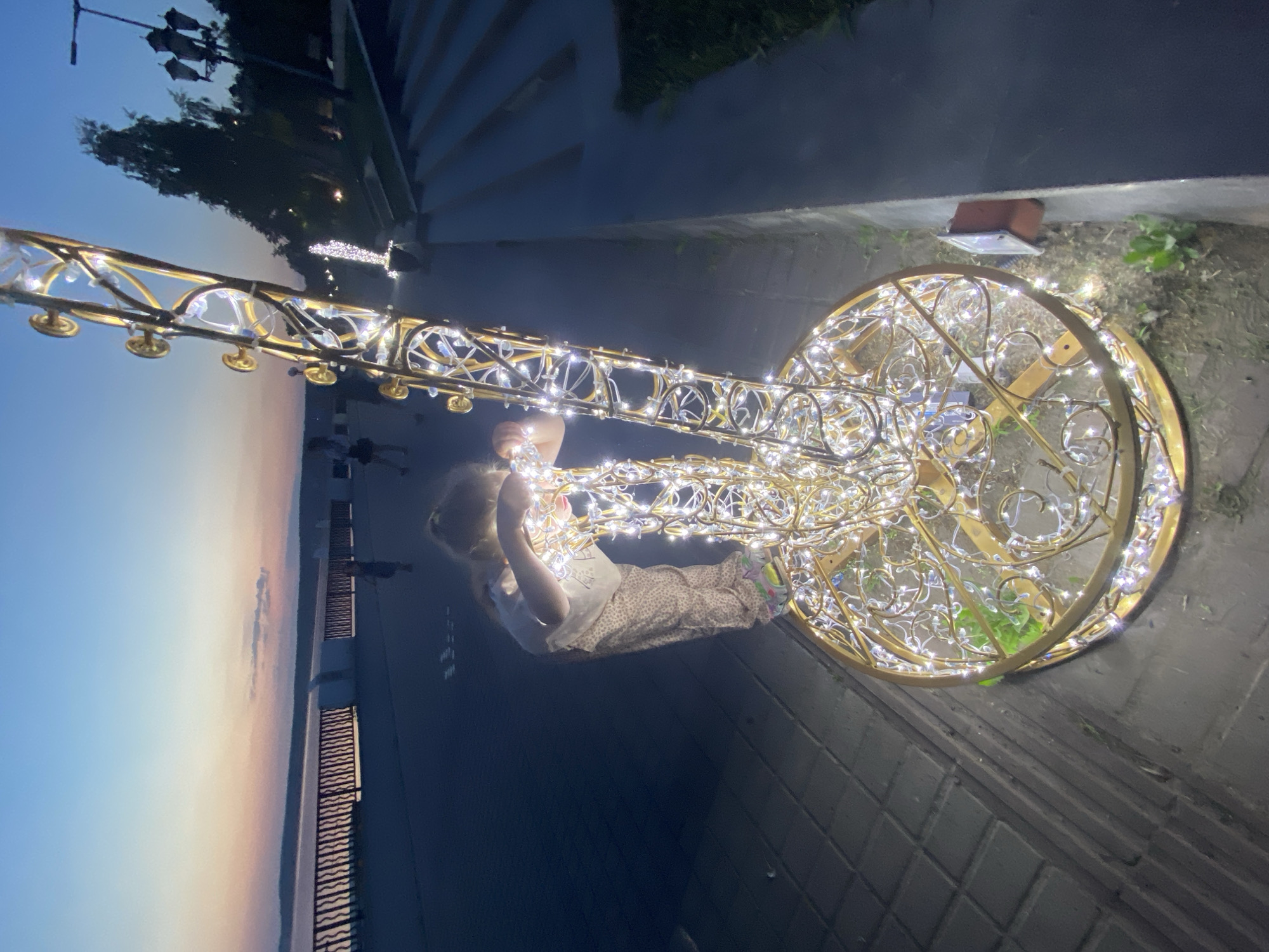 A young child, likely female, is interacting with an illuminated, ornate metal structure in an outdoor setting at night. The structure resembles a saxophone, decorated with numerous small white lights. The child is positioned near the center of the structure, appearing to touch or manipulate the lights. The location appears to be a waterfront area, with a railing and water visible in the background. The scene is near a set of concrete steps and a walkway. Based on the available information, this scene is in Votkinsk, Russia.