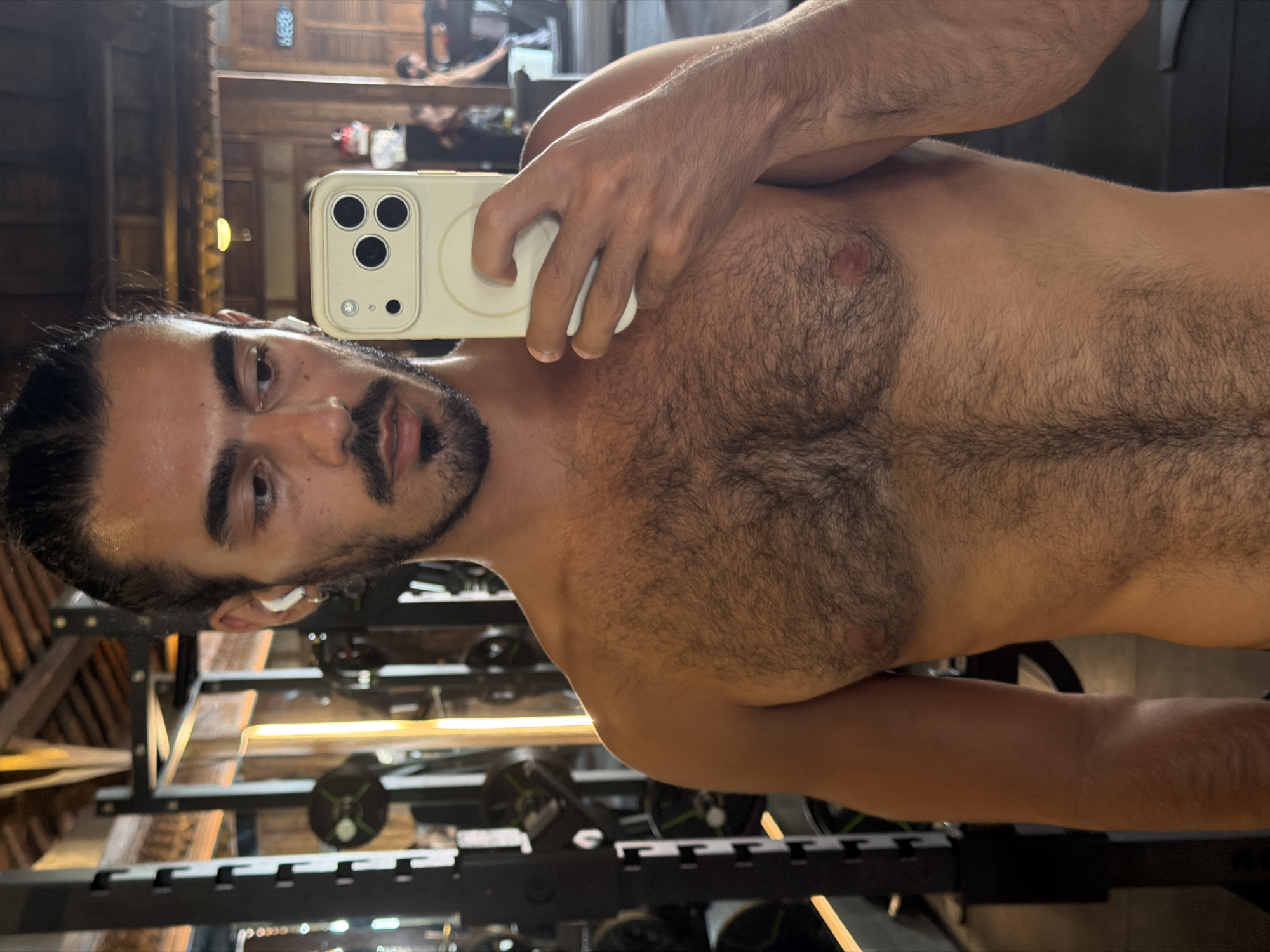 A man with dark hair pulled back, a dark beard, and a light complexion is taking a selfie in a gym. He is shirtless and holding a smartphone with a light-colored case. His left ear is visible with a wireless earbud. He is looking directly at the camera. Behind him is a squat rack with weights. The location is possibly in a gym in North Kuta, Indonesia. In the background, there is a mirror, and a person can be seen reflected in it.