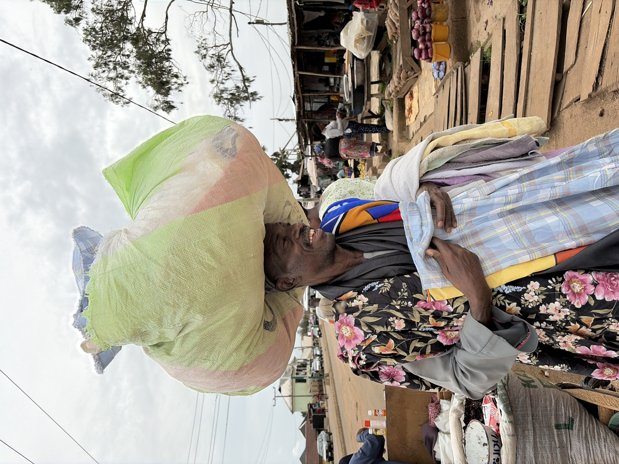 A smiling man, seen from the chest up, is the central subject of this image taken in an outdoor market in Bukuru, Nigeria. He has dark skin and wears a dark shirt with a distinct black and pink floral pattern on the sleeves and shoulders, over a gray undershirt. A large, bulky bundle, wrapped in light green and orange woven material, is balanced on his head, likely containing textiles or clothing. Over his left arm, he carries a neat stack of folded apparel, including plaid shirts and various colored towels.
His wide, joyful smile suggests a friendly disposition or a moment of happiness amidst the activity. The background reveals a bustling market environment with numerous makeshift stalls or shops, characterized by wooden structures and corrugated metal roofs. Other individuals are visible in the background, some sitting, some standing, engaging in market activities. Goods such as baskets of red onions are displayed on the ground, which is partially covered with wooden planks. Power lines stretch across an overcast sky, and some trees are visible in the upper part of the frame. The word "LUXURY" is faintly visible on the cuff of the man's gray shirt sleeve, and indistinct red text can be seen on a white woven sack in the bottom right corner. The scene depicts daytime under a cloudy sky.