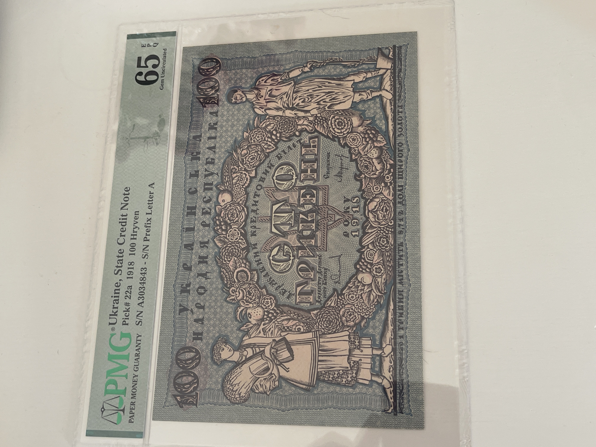An image showcases a Ukrainian 100 Hryven banknote from 1918, professionally graded and encapsulated in a PMG (Paper Money Guaranty) slab. The banknote, identified as Pick# 22a, is graded "65 Gem Uncirculated" and bears the serial number A3034843 with prefix letter A.
The banknote itself features an intricate design in muted blue-grey and reddish-brown tones. Two central figures are depicted: a male figure, likely a worker or farmer, holding an axe or similar tool, and a female figure in traditional attire, carrying a bundle, possibly of wheat. These figures flank an ornate floral wreath that encircles the denomination "100" and the word "ГРИВЕНЬ" (Hryven). The year "1918" is prominently displayed below this central design. Text on the banknote includes "УКРАЇНСЬКА НАРОДНЯ РЕСПУБЛІКА" (Ukrainian People's Republic) and "ДЕРЖАВНИЙ КРЕДИТ" (State Credit). Additionally, a line of text indicates "250 доль щирого золота" (250 parts of pure gold), suggesting a gold backing. The signature "Скарбник" (Treasurer) is also visible.
The banknote is secured within a clear, rigid plastic slab, with a green PMG label visible on the left side, detailing its certification information. The entire piece is laid on a plain, light-colored surface, possibly white or off-white, and captured from an overhead perspective. The image captures the front side of the certified collectible banknote.
