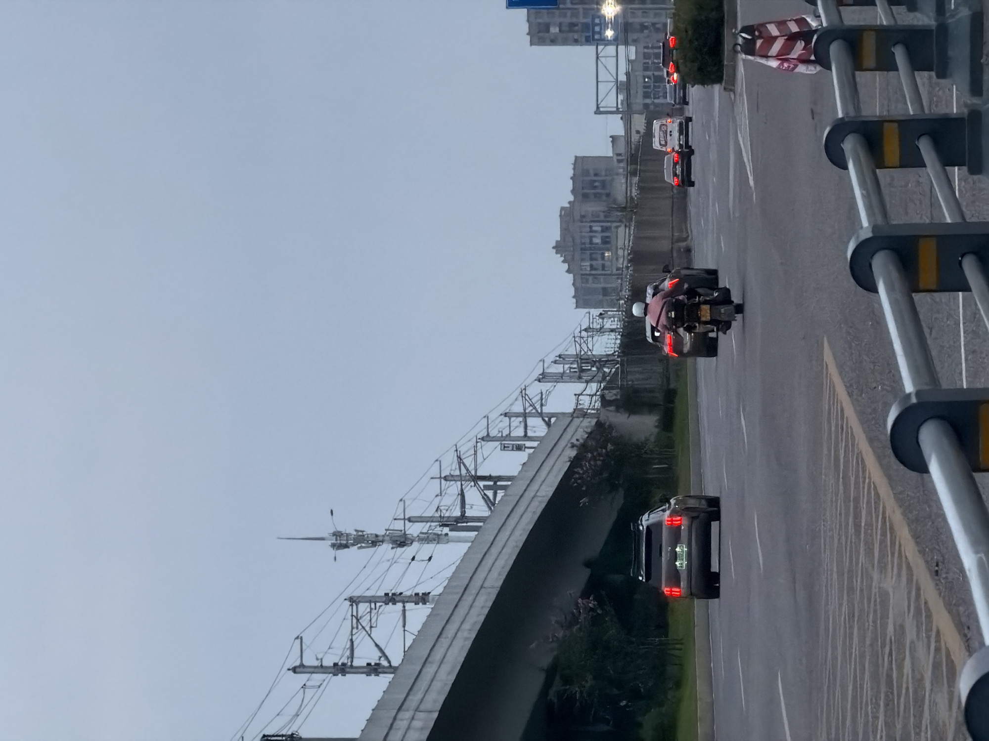 In Jiangjin, China, on an overcast day, a view of a multilane road shows traffic moving beneath an elevated structure carrying overhead railway lines and power lines. On the road, a dark-colored SUV with a roof rack is traveling in the right lane, its brake lights illuminated. To its left, a motorcycle with a rider wearing a pink shirt and helmet is in motion. Further ahead in the distance, other vehicles are visible, including a white van and several cars, navigating the road. The environment is urban, with tall buildings visible in the background beyond the elevated railway structure. A metal railing with orange and black markings runs along the foreground of the road.