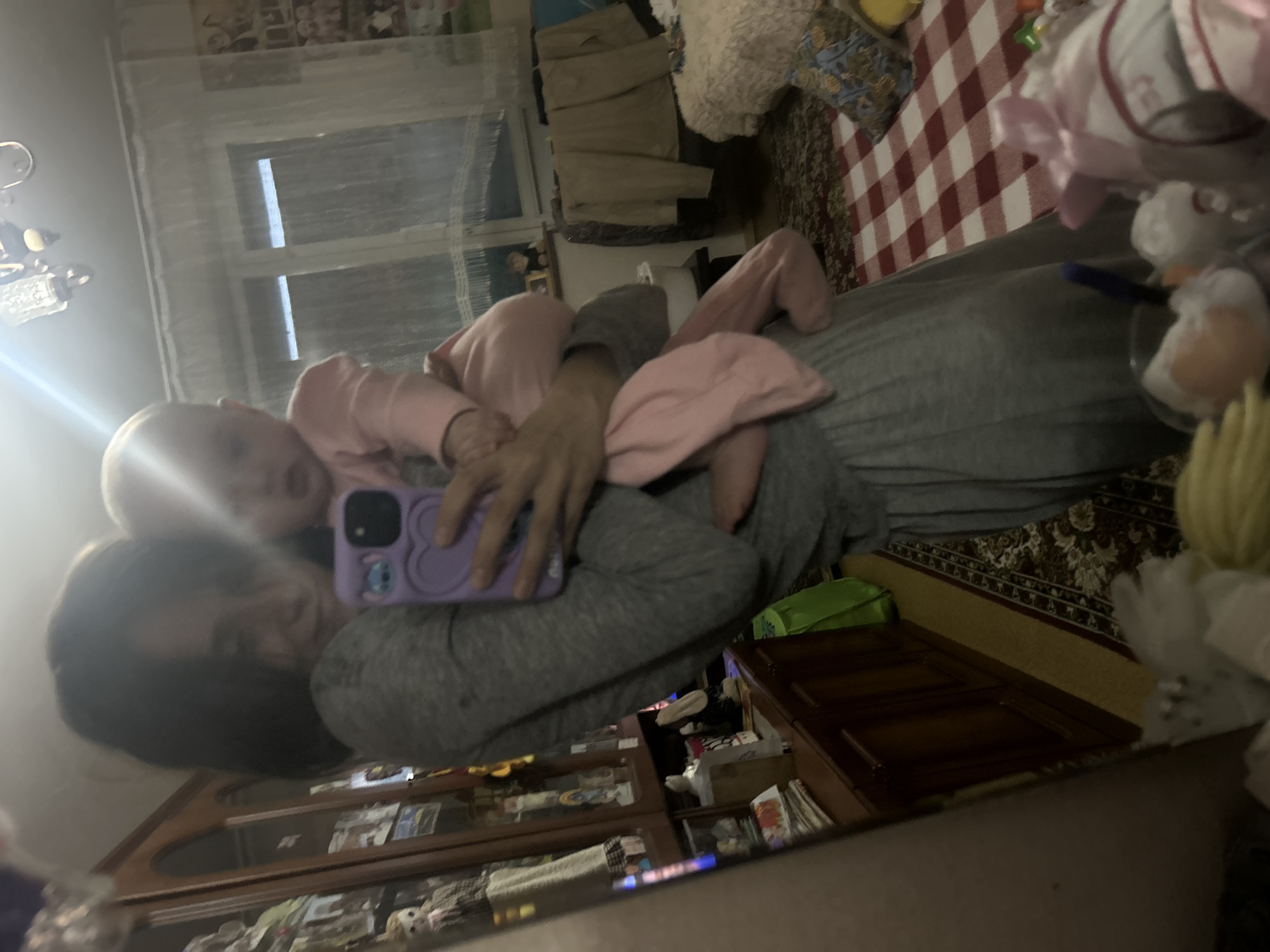 A person, presumably female, is holding an infant in a mirror selfie. The adult wears a gray long-sleeved top, and the baby is dressed in pink. The environment appears to be a domestic setting, possibly a living room or bedroom, suggested by furniture and decorative items. The image is blurry due to the mirror and camera quality. Location information is unavailable.