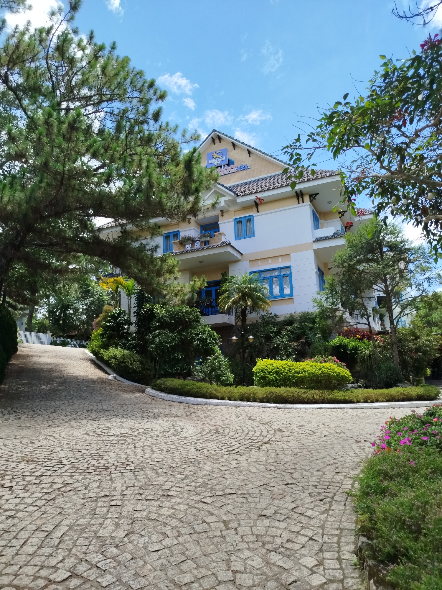 The image shows a three-story building in Lâm Viên, Đà Lạt, Vietnam, characterized by its white walls and blue window frames. A sign with a logo is visible on the roof. Lush greenery surrounds the building, including various trees, bushes, and flowers. The foreground features a cobblestone driveway that curves around a landscaped island with shrubs and flowers. The sky is blue with scattered clouds. No people are visible.