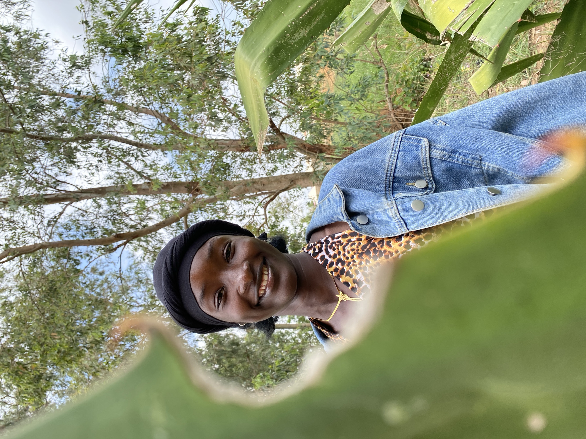 A woman wearing a black headscarf, a leopard-print shirt, and a blue jean jacket is smiling at the camera. She is outdoors with trees and foliage visible in the background. The location is not specified as being in Durimi, Nigeria. Large, green, fleshy plant leaves partially obscure the foreground. A gold necklace with a star pendant is visible on the woman's neck.