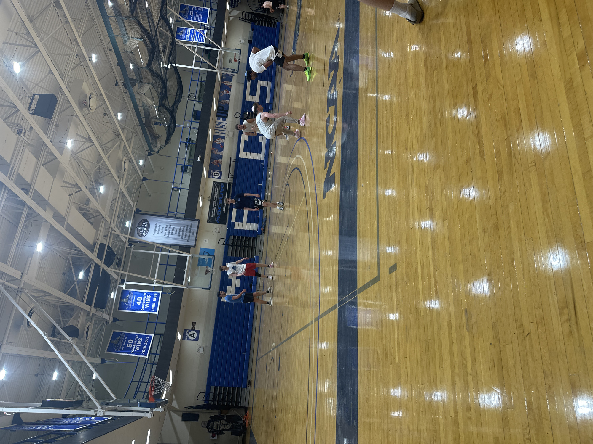 Here is a description of the image:
The image depicts an indoor basketball court, likely located in a school or university gymnasium, possibly in the city of New York in the United States of America. The court has a wooden floor with blue painted lines and the word "NCAA" printed in blue. There are several people on the court, some of whom appear to be stretching or warming up. There are blue bleachers along one side of the court. Banners and signage are hanging from the walls. One banner celebrates "50 Consecutive Wins 2019-2022," and another mentions "40 Consecutive Wins 1995-1999" suggesting a successful sports program. The gym has a high ceiling with lights.