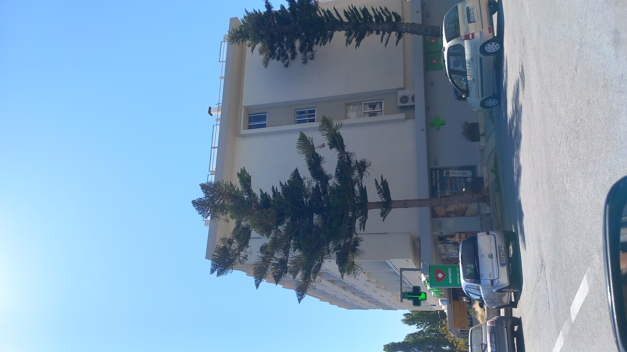 The image depicts a street scene in the city of Kos, Greece, on a clear, sunny day. In the background, a modern, multi-story building with a light-colored facade dominates the frame. The building has several windows and a balcony railing on the top floor. In front of the building, a green pharmacy cross sign is visible, along with storefronts. One storefront displays advertisements for "FREZYDERM" and "GOVELVET". There are two trees with distinctive needle-like foliage positioned in front of the building.
In the foreground, the street is paved with asphalt, and white lane markings are visible. Several cars are parked or driving on the street. A silver hatchback with a license plate ending in "4371" is parked in front of the pharmacy. To its left, a smaller, darker colored car with a license plate ending in "6159" is also visible. A third silver hatchback with a license plate ending in "7613" is parked on the right side of the frame. Part of a vehicle's side mirror is visible in the bottom corner of the image, suggesting the photo was taken from inside a car. The overall environment appears to be a commercial or urban area.