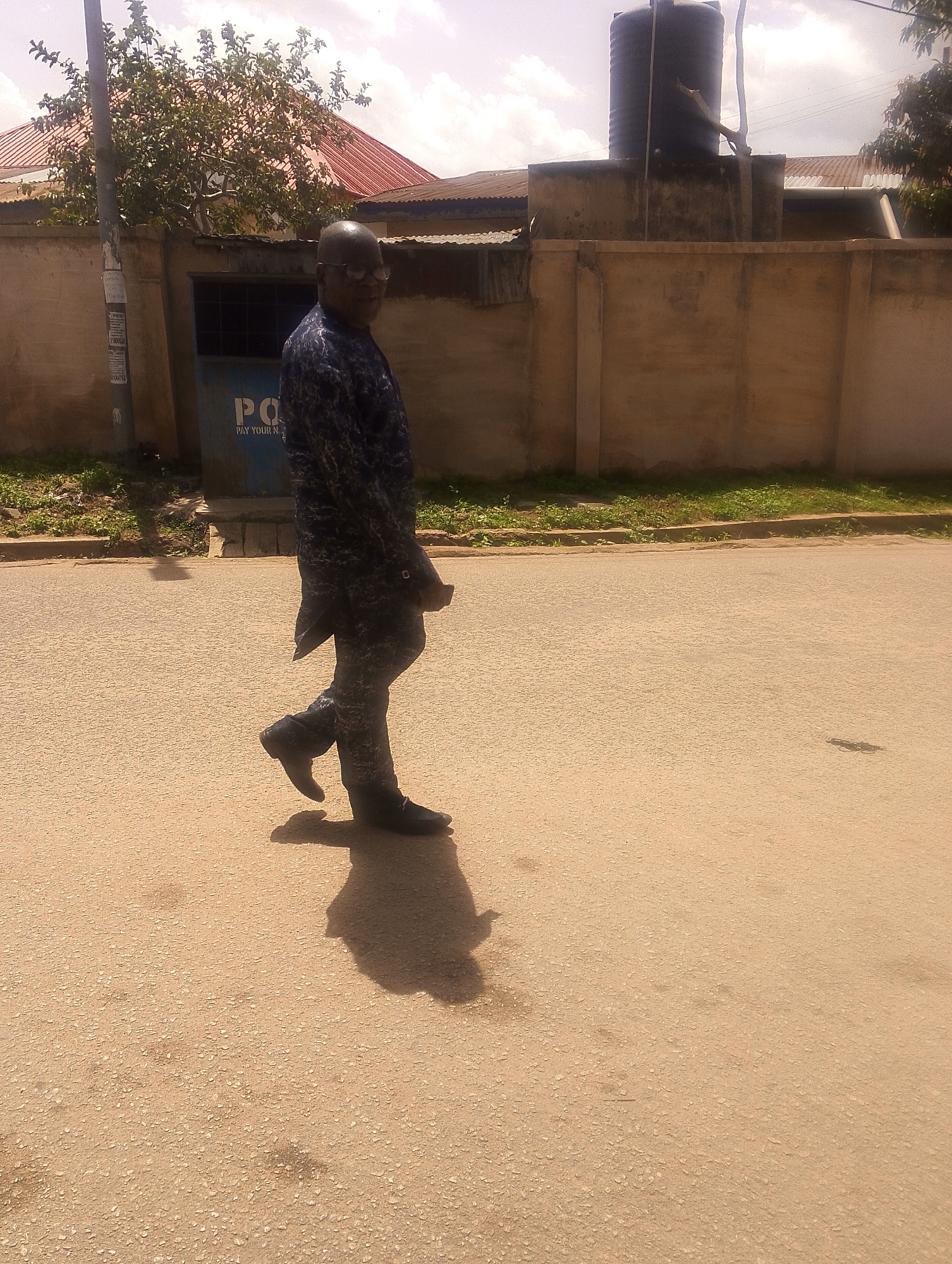 This media file depicts a man walking on a paved road in Jos, Nigeria. The man is wearing a dark, patterned shirt and pants, and dark shoes. He is walking from left to right in the frame, facing away from the camera, with his head turned to look back over his shoulder. He appears to be of average build.
The setting is an outdoor environment, likely a street or sidewalk. In the background, there is a building with a red-tiled roof and a tan-colored wall. A large, black water tank is visible on top of a concrete structure behind the wall. A pole with some signage is also present on the left side of the frame. Some greenery, including trees and shrubs, can be seen growing along the wall and the edge of the road. The road surface is asphalt and appears to be a light brown or tan color.
The time of day appears to be daytime, with sunlight casting shadows. The sky is visible in the upper portion of the image and appears partly cloudy. The overall impression of the scene is one of everyday life in an urban or semi-urban setting.
There is some visible text on a blue object behind the man, which appears to be part of a payment or utility kiosk. The visible text includes "PO" and "PAY YOUR N". The rest of the text is not clearly legible.
