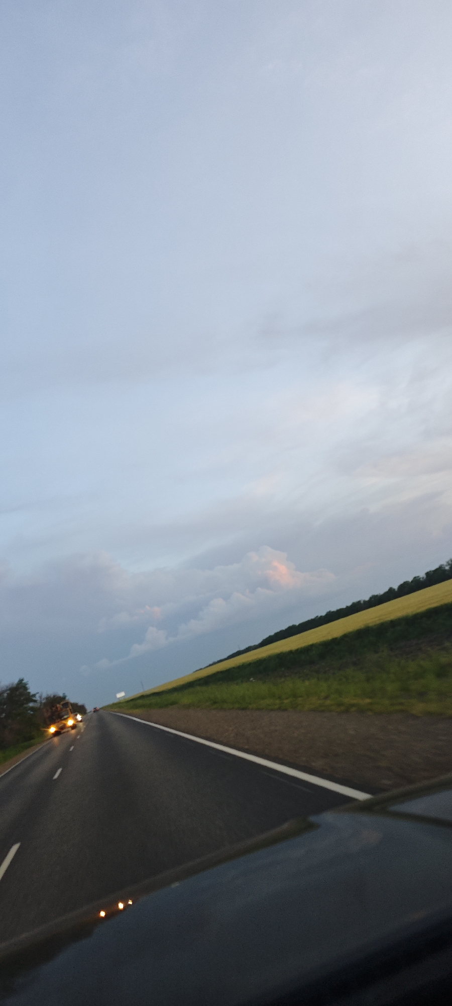 The image shows a two-lane road in a rural setting, possibly in the vicinity of Beysug, Russia. The road is asphalt, with a white line marking the edge. A truck with illuminated headlights is approaching in the opposite lane. To the right of the road, there is a sloping landscape with green vegetation and a field of yellow crops. The sky is overcast with clouds. The photograph appears to have been taken from inside a vehicle, as parts of the car are visible in the foreground.
