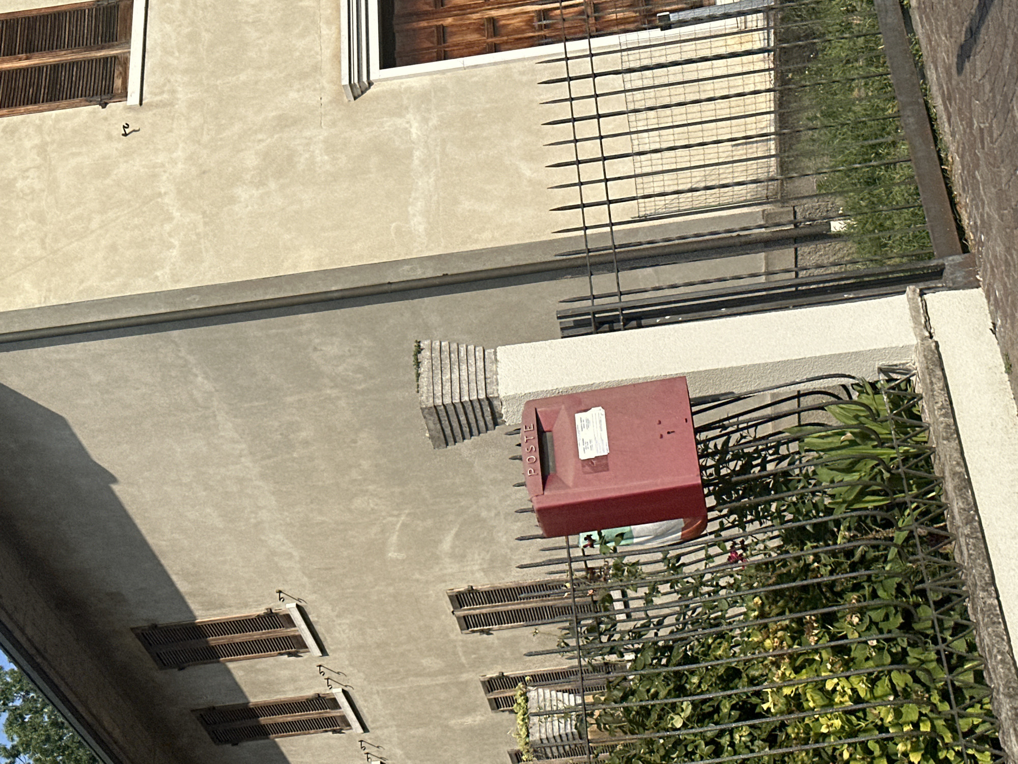 The image captures a sunlit exterior scene in Rudiano, Italy. A weathered, beige building with dark-slatted windows is the primary structure. A bright red "POSTE" mailbox stands on a concrete post to the left of a dark metal fence that encloses a patch of lush green vegetation. The fence runs horizontally across the foreground of the image. A sliver of a wooden door is visible on the right side of the building. The ground is paved with cobblestones. The lighting suggests it is daytime, with shadows cast by the building and fence.