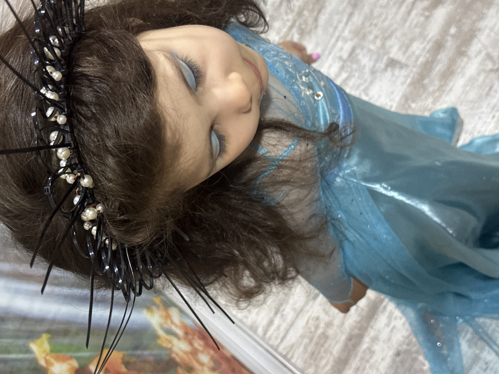 A young child lies on a textured surface, possibly a bed or floor, wearing a light blue dress with glitter accents. The child has dark hair and is adorned with a black crown featuring white pearl-like embellishments and extended black spikes. The child's eyes are closed and are coated with light blue eyeshadow. The background includes a hint of artwork with autumnal colors, possibly indicating a residential setting.