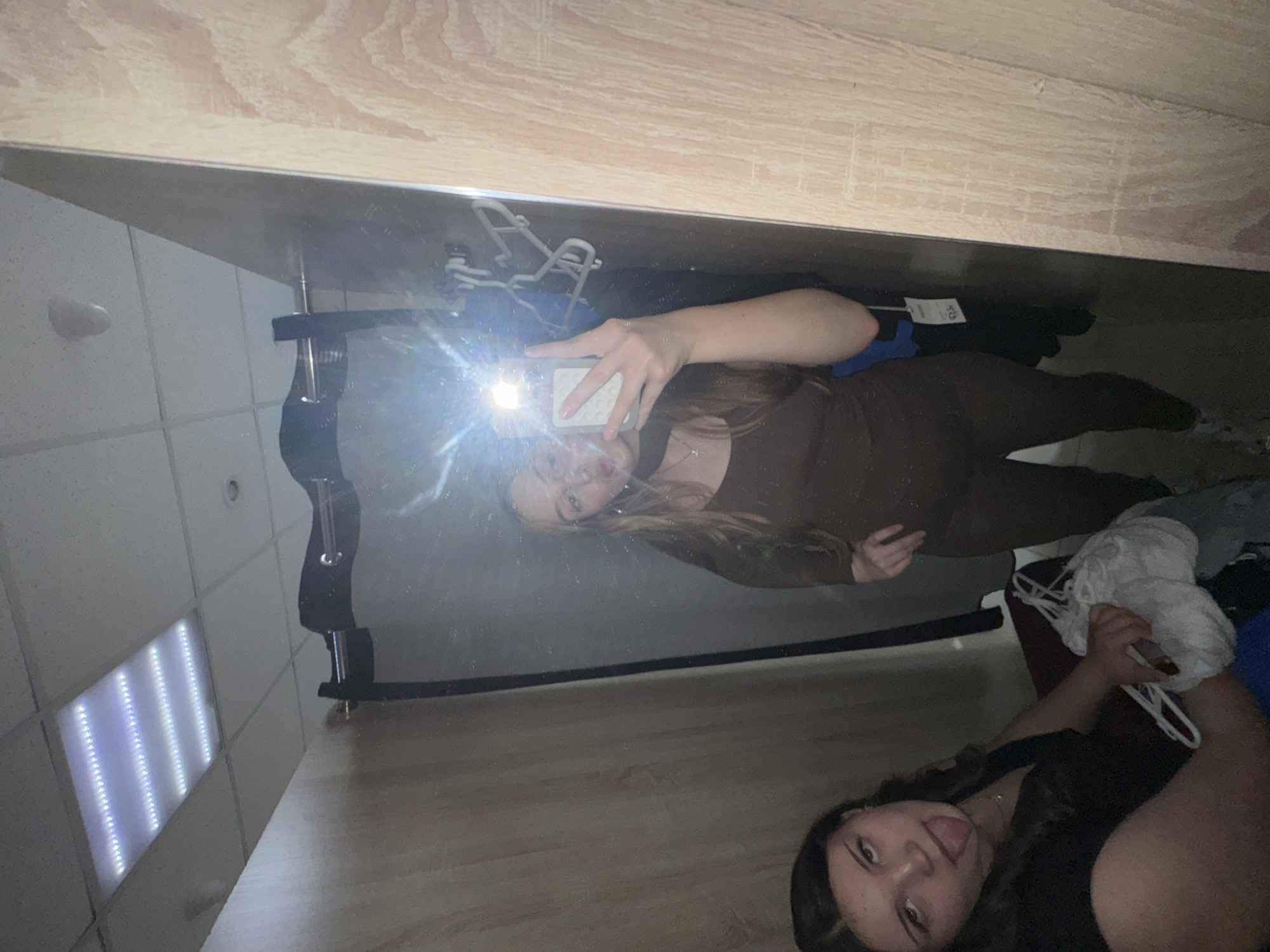 Two young women are in a dimly lit fitting room. On the left, a woman with dark hair is leaning forward, sticking her tongue out at the camera, and holding a bundle of white clothing. On the right, reflected in a mirror, a woman with long blonde hair is taking a selfie, making a peace sign with her hand. The fitting room has wooden paneled walls and a tiled ceiling with fluorescent lights. There are hangers visible in the mirror, along with a dark curtain. The image appears to be a candid moment captured in a public fitting room. No specific location within the city of Perm, Russia, can be identified from the image.