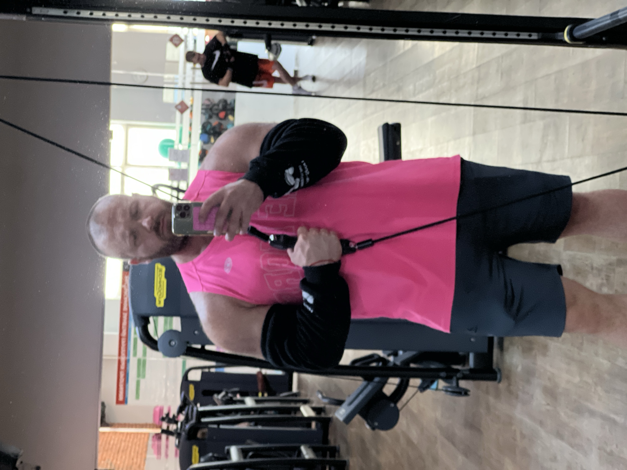 A bald, white male with a beard, wearing a bright pink tank top and dark shorts, stands in a gym and takes a selfie in a mirror. He is holding a cable machine handle in his right hand and his smartphone in his left. Black arm sleeves are worn over his biceps. In the background, other gym equipment is visible, including weight machines and a rack of dumbbells. A second man in a black t-shirt and orange shorts can be seen in the mirror's reflection, using gym equipment. The gym floor is light-colored wood, and the walls are white with various posters and signs. There are large windows in the background, suggesting natural light entering the space. The overall environment appears to be a functional fitness facility.