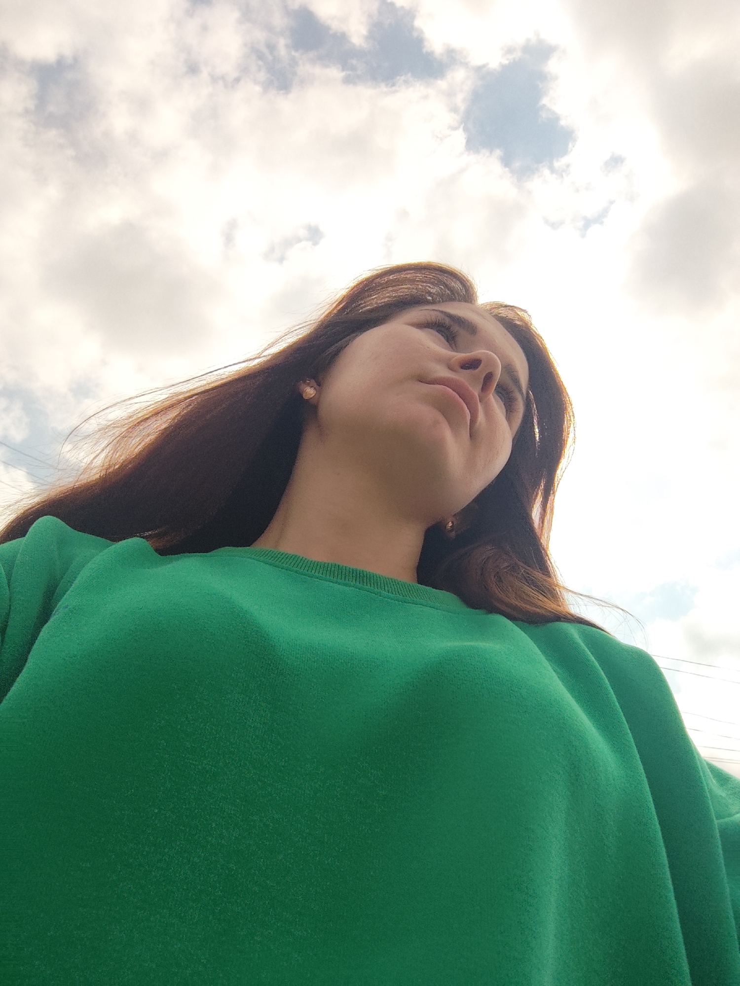 A low-angle shot captures a young woman looking up at the sky. She has long, dark brown hair that falls around her shoulders and is wearing a bright green crewneck sweatshirt. The background is dominated by a cloudy sky with patches of blue visible. The scene appears to be outdoors in a non-descript urban or suburban environment, with faint power lines visible in the distance. There is no indication of the specific location within Фоки, Russia.