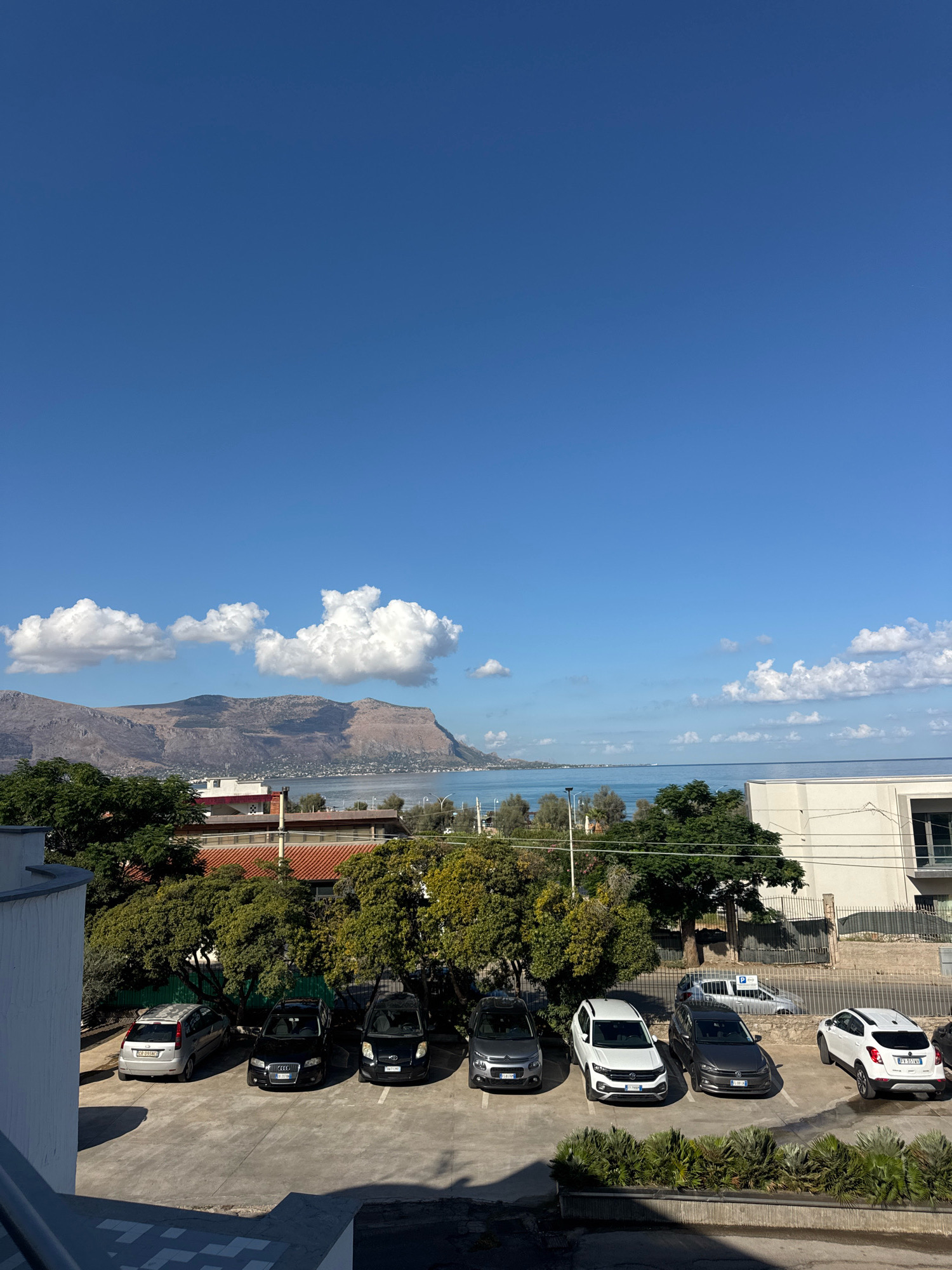 The image captures a sunlit scene in Isola delle Femmine, Italy, featuring a panoramic view of the coastline and mountains under a clear blue sky with scattered white clouds. In the foreground, a parking lot is filled with several cars including a silver Ford Fiesta, a black Audi A4, a black Fiat Panda, a gray Citroen C3, a white Volkswagen T-Roc, a gray Volkswagen Polo, and a white Fiat 500X. Lush green trees are interspersed throughout the scene, with some obscuring the view of buildings behind them. The buildings appear to be residential or commercial structures, with tiled roofs visible. In the background, a calm sea meets a rugged, mountainous coastline, suggesting a coastal town setting. The overall atmosphere is one of a typical sunny day in a Mediterranean locale.