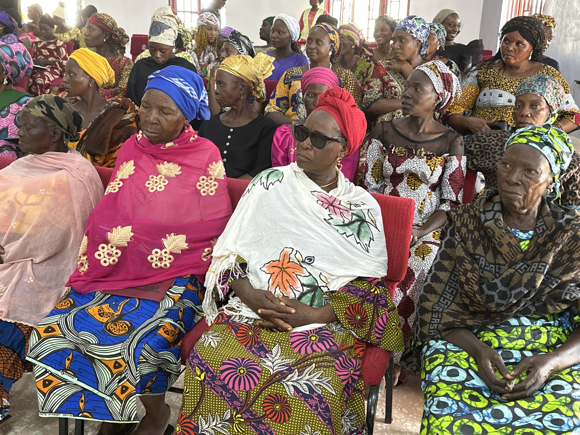 This image depicts an indoor gathering of approximately 20-30 adult women, predominantly older, seated in rows within a community hall or similar venue in Goni Gara, Nigeria. The women are uniformly dressed in vibrant, patterned traditional African fabrics, with many wearing colorful headwraps or scarves.
In the foreground, from left to right:
1. A woman wearing a camouflage-patterned headwrap and a light pink garment.
2. A woman with a blue headwrap, dressed in a bright pink top adorned with gold embroidered leaf and circular patterns, over a blue skirt featuring orange and white wave and geometric designs.
3. A woman wearing a red headwrap and black sunglasses, with gold heart-shaped earrings. She is wrapped in a white shawl with large painted orange, green, and pink floral motifs, over a green skirt with intricate pink, red, and white flower patterns. Her hands are clasped in her lap.
4. An older woman with a green and blue patterned headwrap, draped in a dark brown and black geometric patterned shawl, worn over a bright green skirt with blue and black circular designs. Her hands are clasped, holding a small, indistinct object.
The background reveals additional women, similarly attired in diverse patterned fabrics and head coverings, seated behind the foreground individuals. Most women maintain neutral or contemplative expressions, generally looking forward or slightly to the side, with one woman in the mid-ground smiling. The seating consists of red chairs or benches arranged in an audience-like configuration. The walls are light-colored, and natural light enters through windows visible in the upper background. The floor is light-toned.