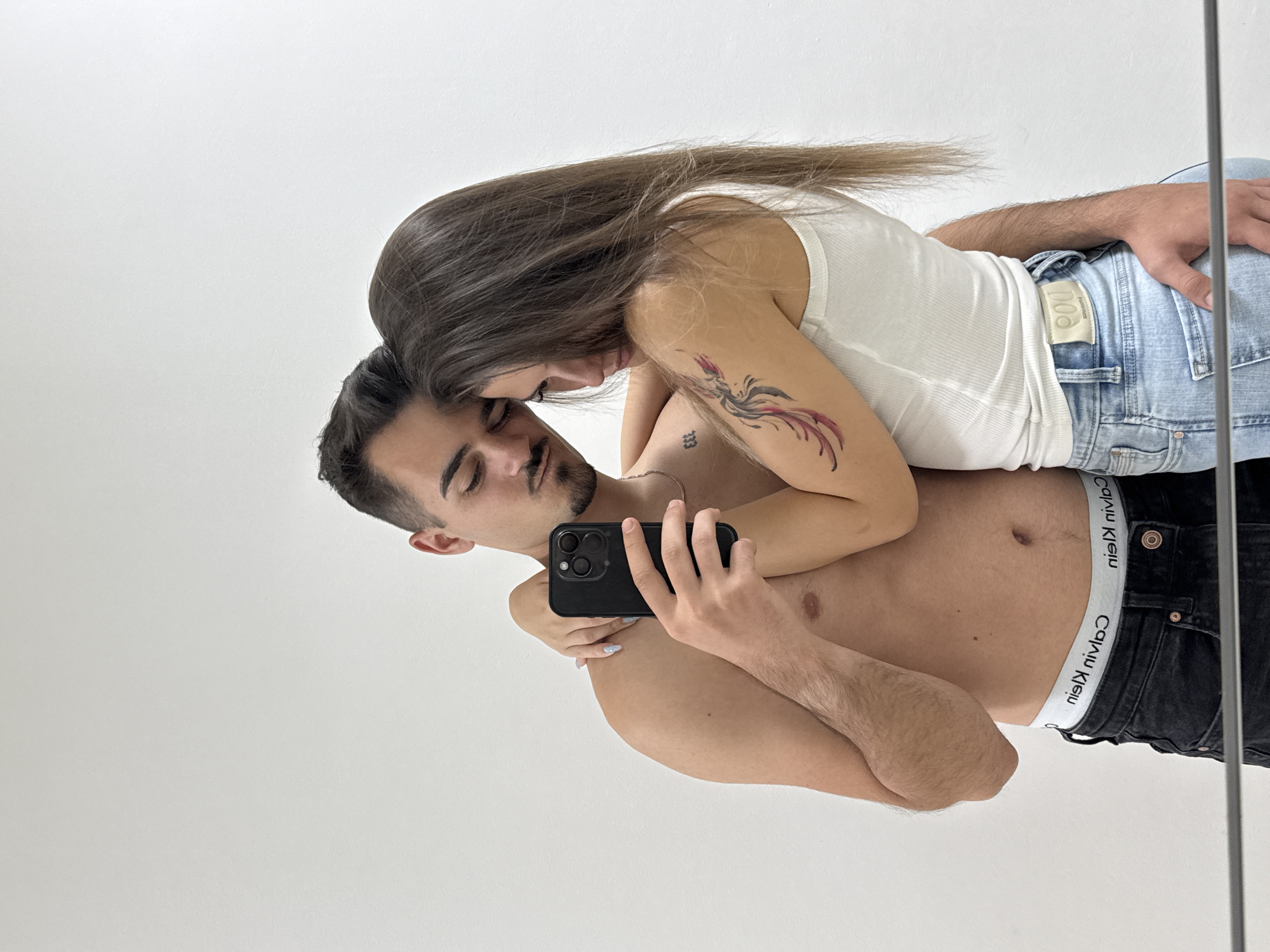 Two individuals, a male and a female, are captured in a mirror selfie, standing in a close embrace against a plain white background in Sofia, Bulgaria. The male, shirtless, with a fade haircut, mustache, and short beard, wears black jeans and a visible white "Calvin Klein" underwear waistband (text appears reversed as "niel nivlaC" due to reflection). A small "333" tattoo is present on his left rib cage, and a thin gold-colored chain is around his neck. He holds a black smartphone with a triple-lens camera in his right hand, pointed towards them. His left arm is around the female's back. The female, with long dark brown hair, wears a white ribbed tank top and light blue denim jeans. A colorful tattoo resembling a phoenix is visible on her left upper arm. Her right arm is around the male's neck/shoulder, with light-colored fingernail polish visible, and her left hand is placed on his lower back/waist. Their heads are close together, and both appear to be looking downwards. A dark horizontal bar, indicative of a reflective surface, is visible at the bottom of the frame.