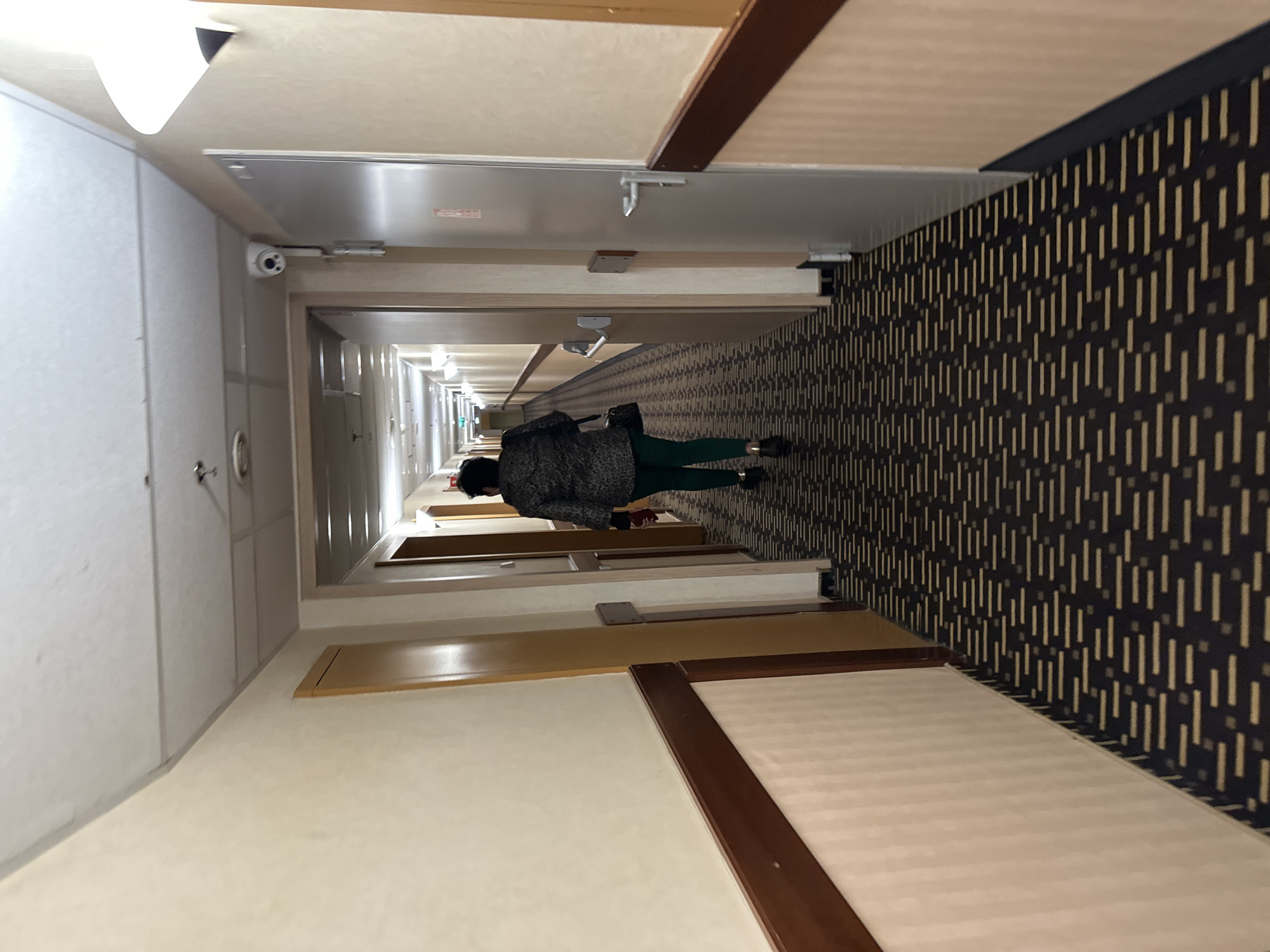 The image shows a person walking down a hotel hallway. The hallway has beige walls with dark brown trim around the door frames. The doors themselves are light beige with a silver door. The floor is a patterned, black and gold rug. The ceiling has recessed lighting. The person is wearing a dark jacket with a leopard print and green pants. They are walking away from the camera down the hallway. There is a security camera mounted on the ceiling near the beginning of the hallway. This image was taken in Wrocław, Poland.