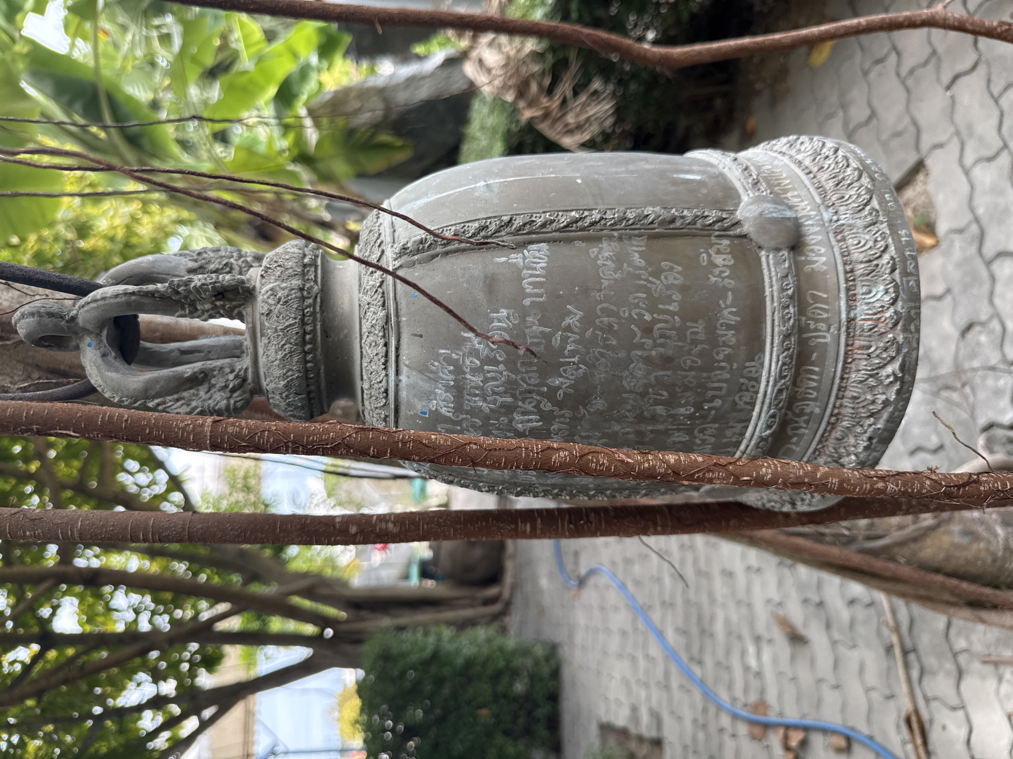 The image shows a close-up of a large, weathered, bronze bell hanging from tree roots. The bell is adorned with intricate carvings and inscriptions in Thai script. The background is a blurred outdoor scene with trees, foliage, and a paved walkway. A blue hose is visible on the walkway. The setting appears to be a temple or a garden in Tambon Na Chom Thian, Thailand, suggested by the cultural elements and the typical vegetation of the region. There are no people visible in the image. The lighting suggests it is daytime.